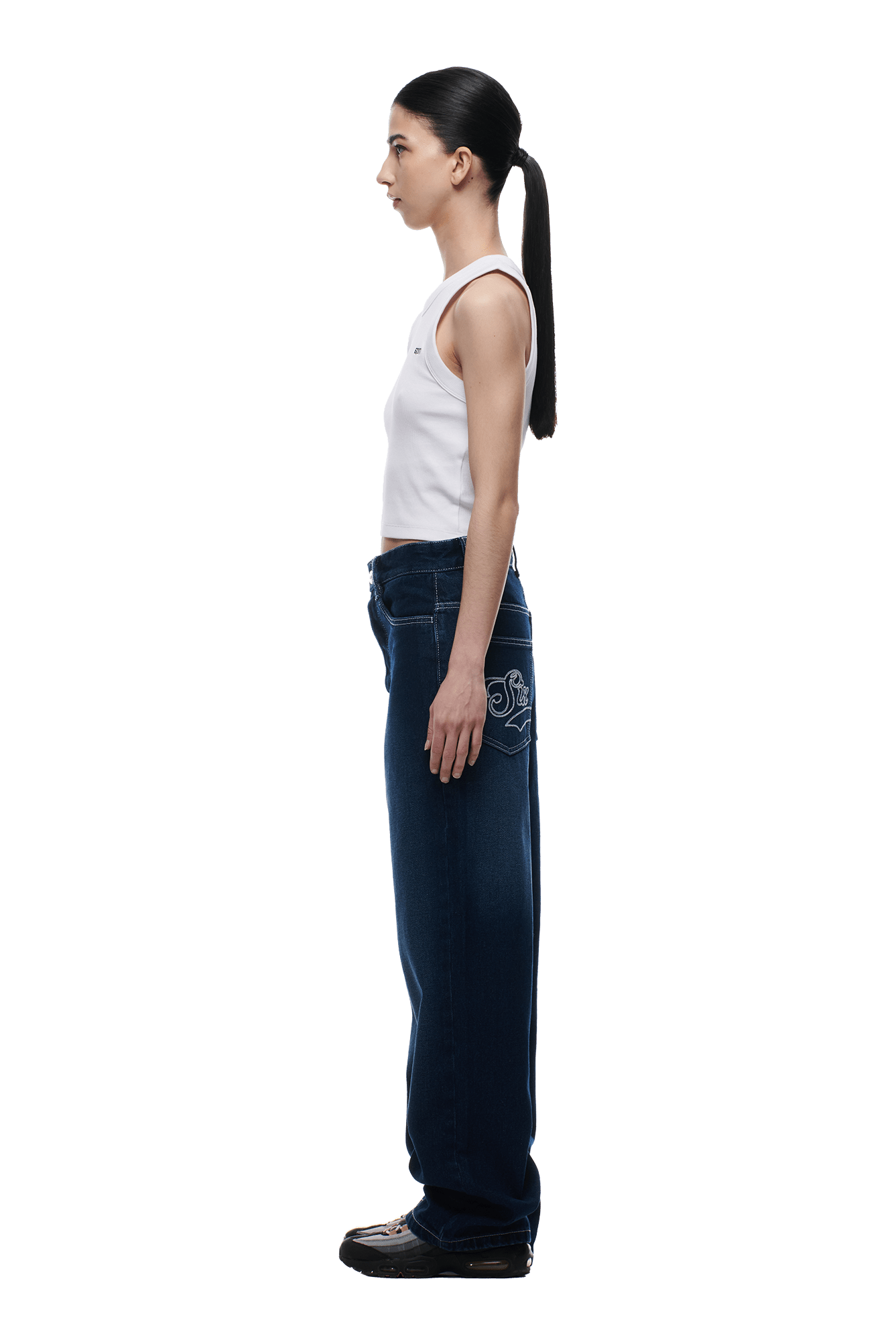 A woman stands in profile against a white background, wearing a white sleeveless top, 6PM SCRIPT LOGO DENIM WASHED DARK BLUE baggy jeans with back pocket embroidery, black shoes, and her hair tied back in a long ponytail.