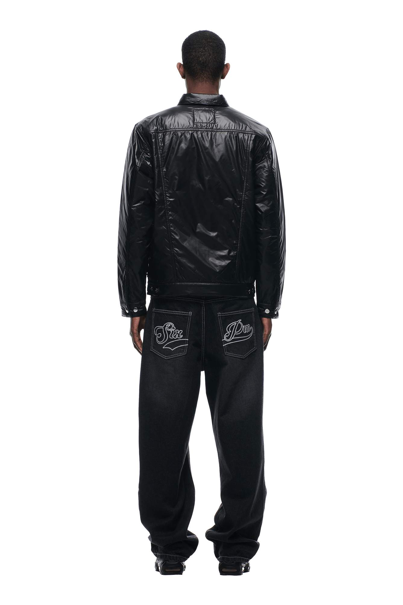 A person faces away, wearing the 6PM PADDED TRUCKER JACKET BLACK and loose black jeans with large embroidered designs on the back pockets, against a plain white background.