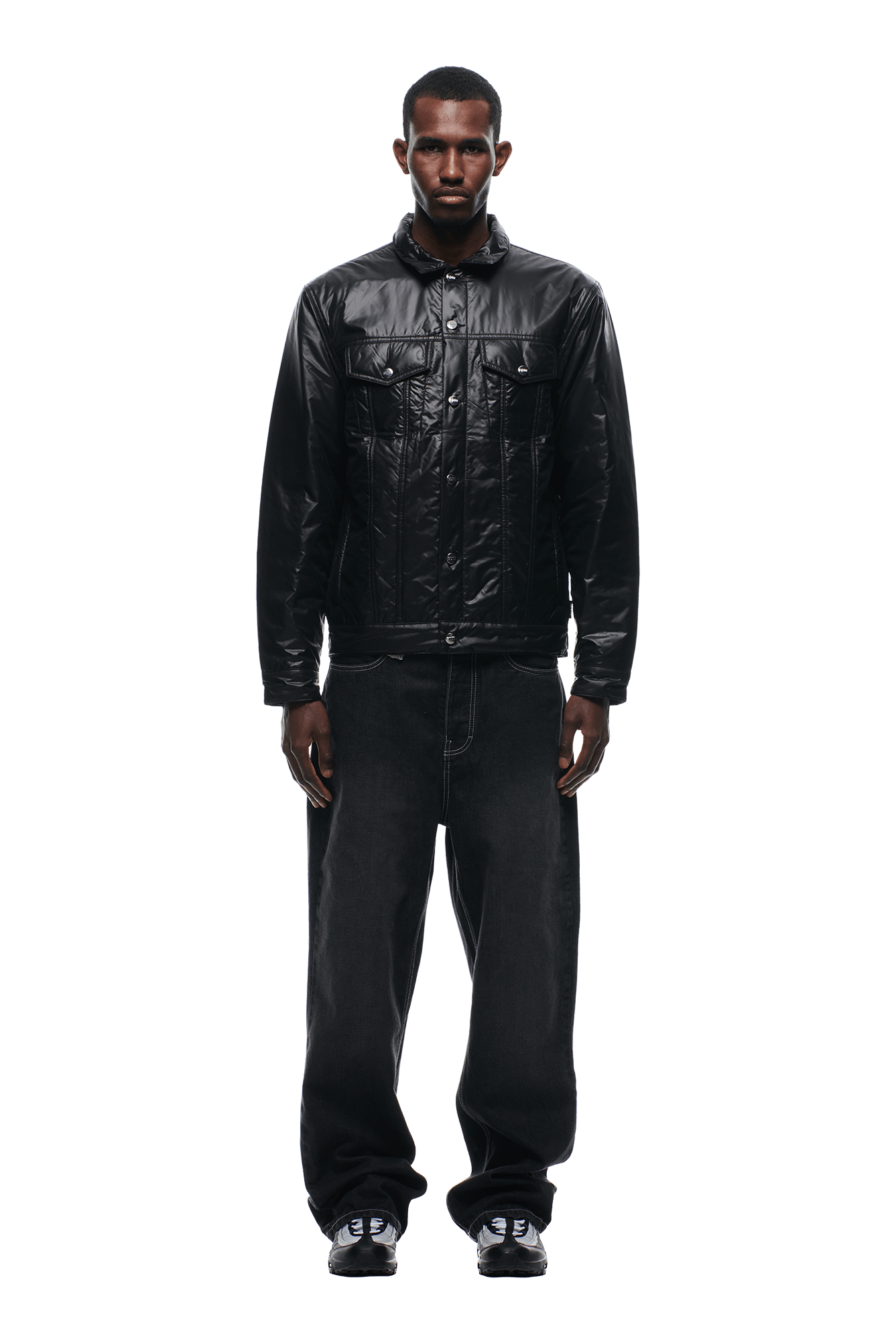 A man faces forward against a plain white background, wearing the 6PM PADDED TRUCKER JACKET BLACK, paired with baggy black jeans and black sneakers.