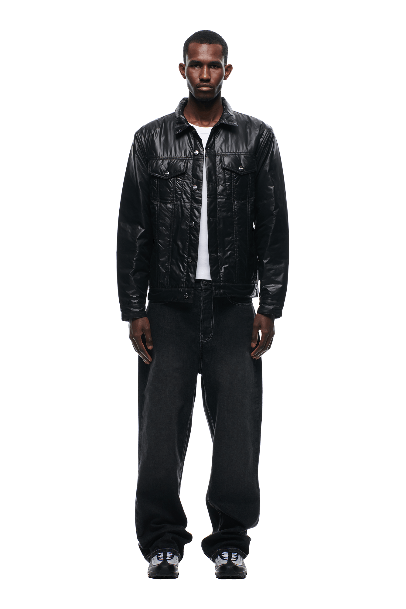 A man faces forward against a white background, wearing the 6PM PADDED TRUCKER JACKET BLACK over a white shirt, paired with loose black jeans and black sneakers.