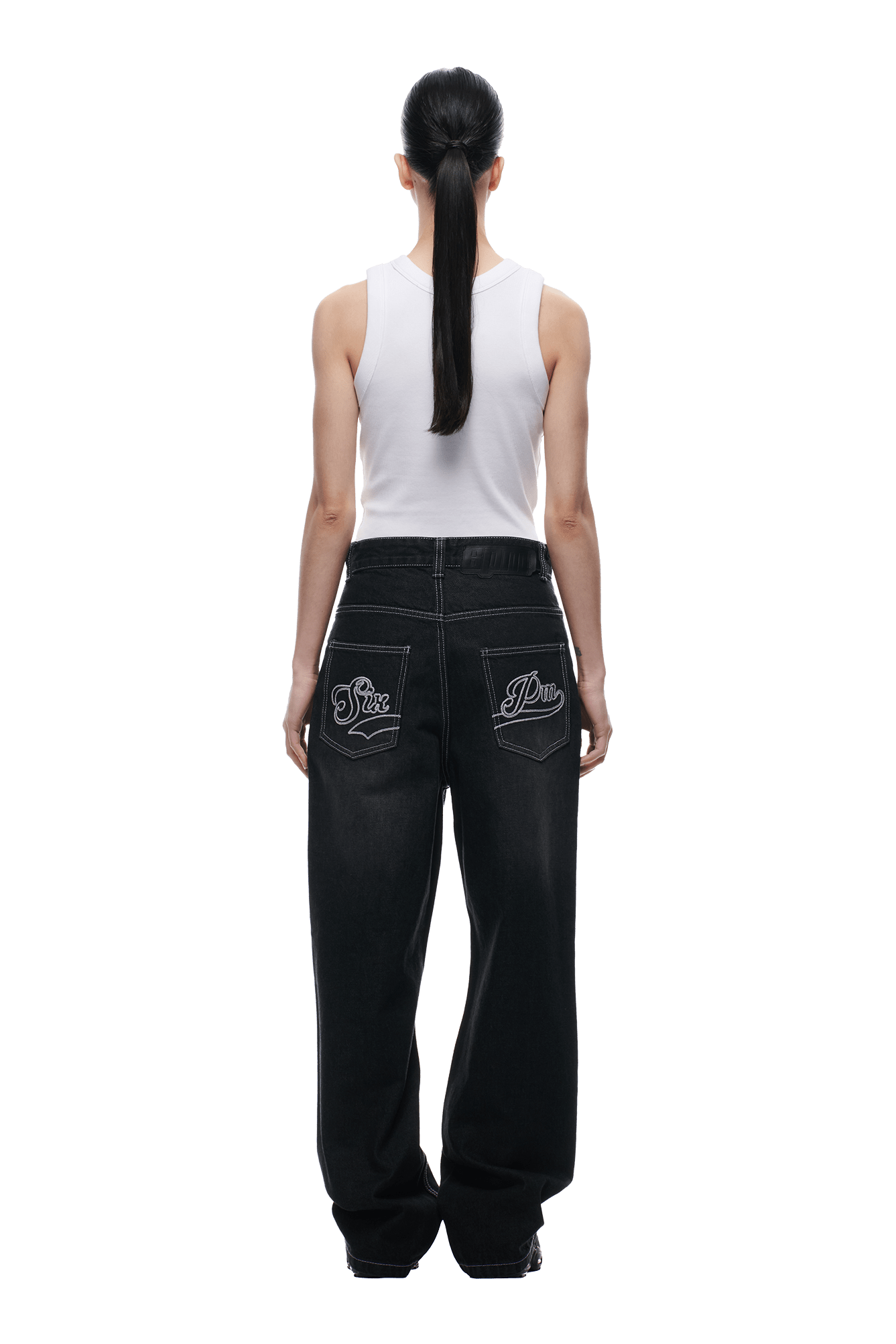 A person with long dark hair in a ponytail wears a white sleeveless top and 6PM SCRIPT LOGO DENIM WASHED BLACK jeans with white stitching and embroidered back pocket logos, standing against a plain white background, shown from behind.