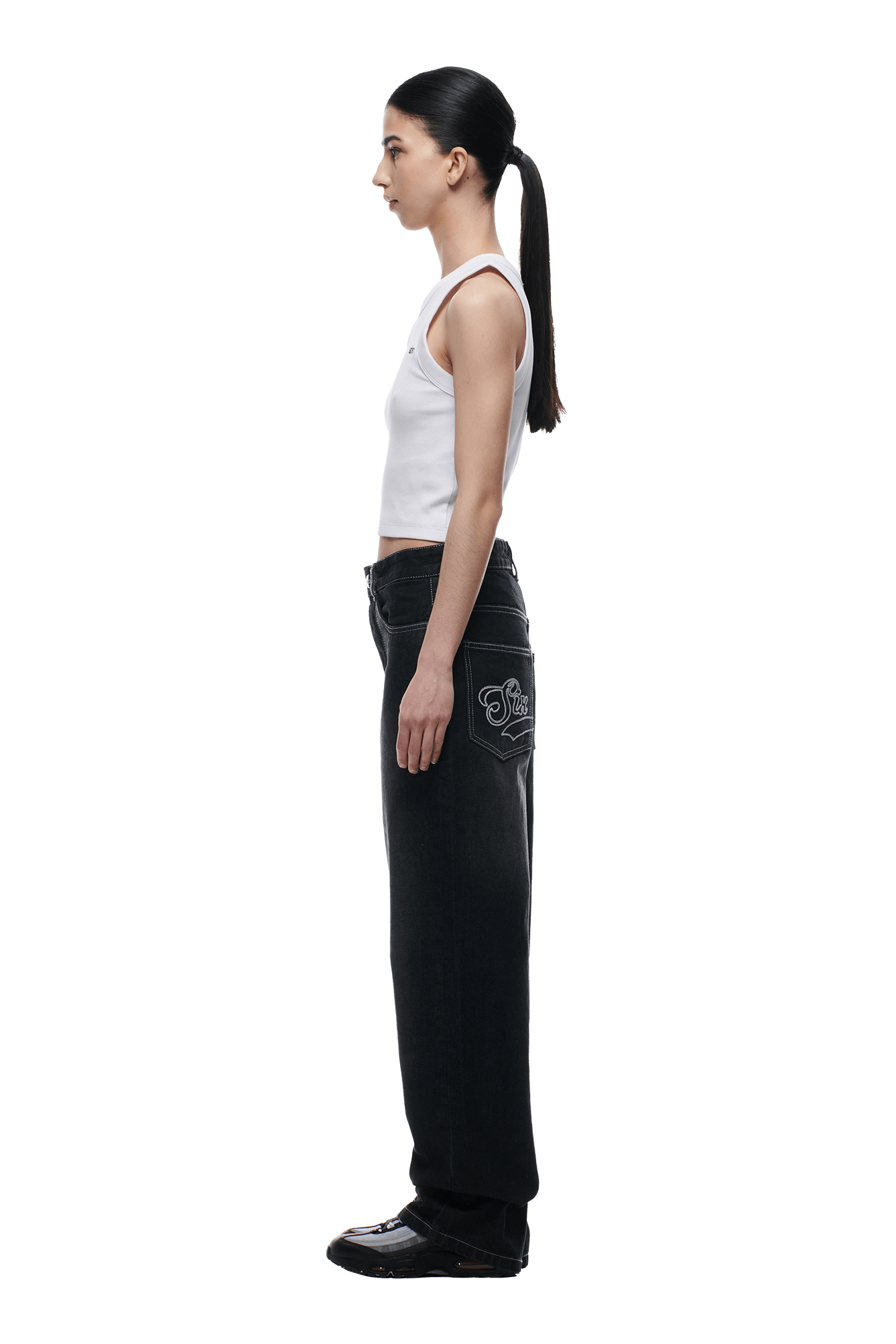 A woman stands in profile against a white background, wearing a white sleeveless top, 6PM SCRIPT LOGO DENIM WASHED BLACK wide-leg jeans with a graphic back pocket, black shoes, and her dark hair tied back in a low ponytail.