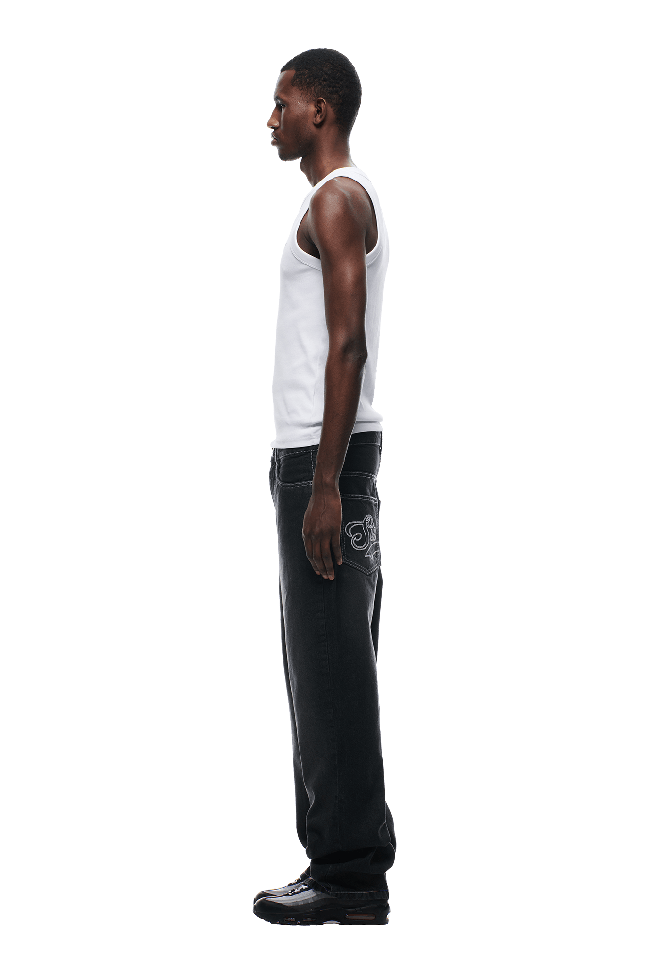 A man in profile against a white background wears a white tank top, 6PM SCRIPT LOGO DENIM WASHED BLACK jeans featuring white embroidery on the back pocket, and black shoes.