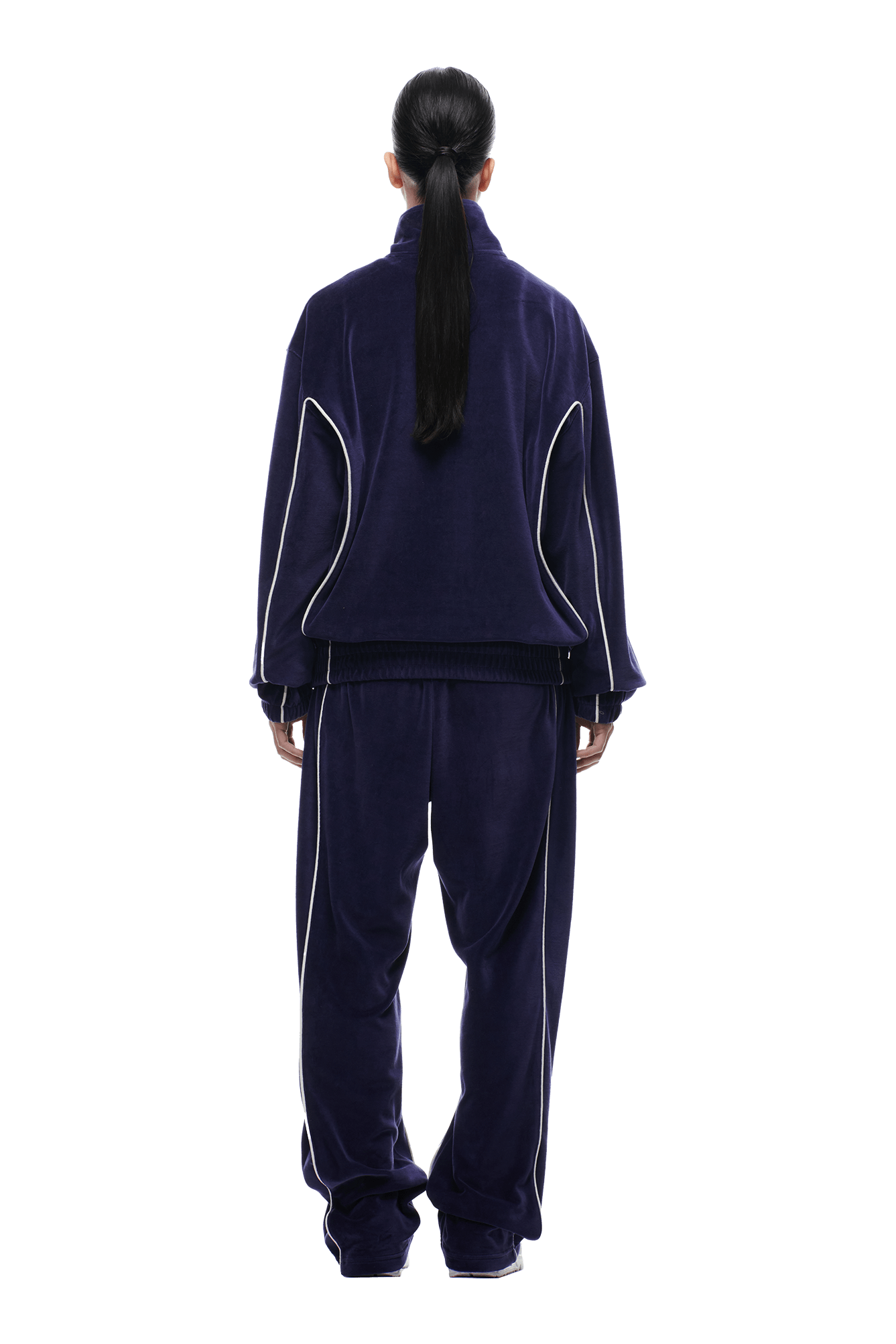 A person with long dark hair in a ponytail stands with their back to the camera, wearing the 6PM VELOURS TRACK JACKET ORBIT BLUE. The jacket is loose-fitting with white piping, set against a plain white background.