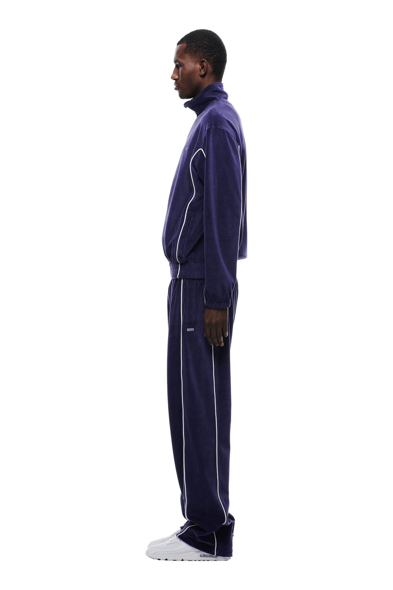 A man stands in profile against a plain white background, wearing the 6PM VELOURS TRACK JACKET ORBIT BLUE with white piping and white sneakers.