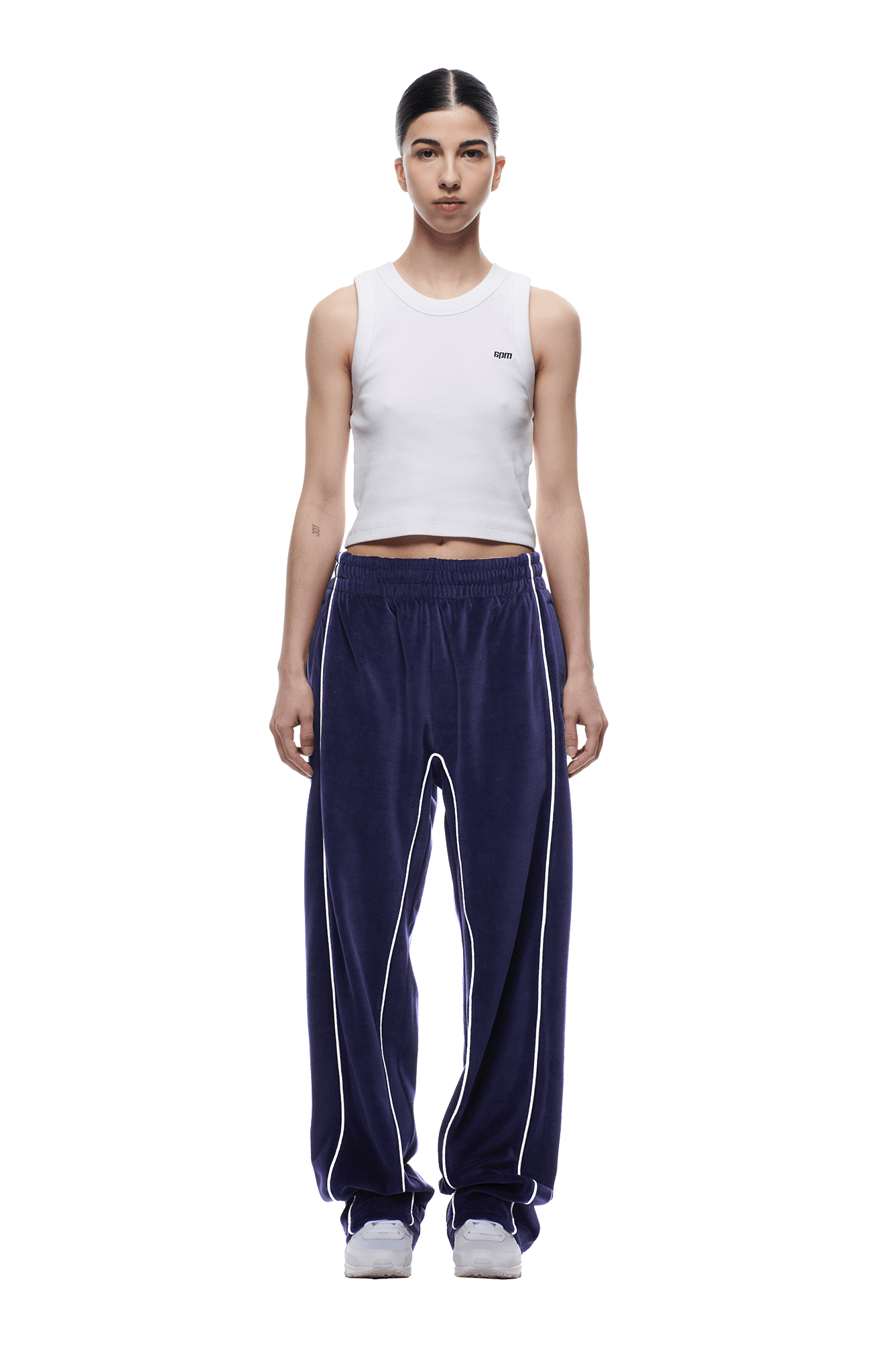 A person faces forward against a white background, wearing the 6PM VELOURS TRACK PANT ORBIT BLUE with white piping, paired with a white sleeveless crop top and white sneakers.