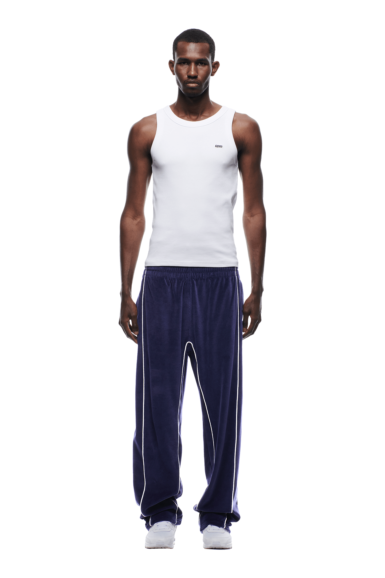 A man stands facing forward against a white background, wearing 6PM VELOURS TRACK PANT ORBIT BLUE with white piping, a fitted white tank top, and white sneakers.