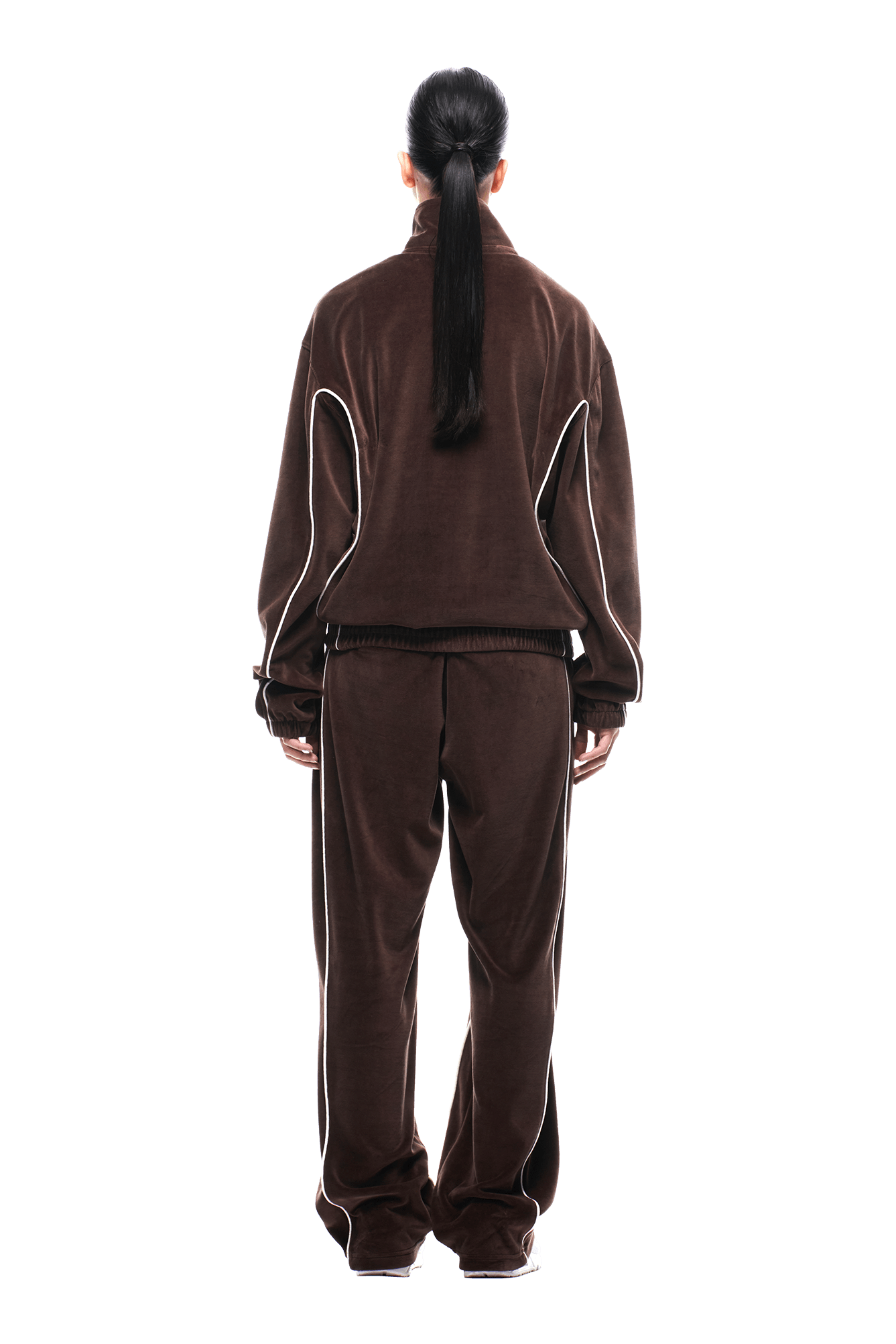 A person stands facing away against a plain white background, wearing the 6PM VELOURS TRACK JACKET CHOCOLATE BROWN, a loose-fitting jacket with white piping on the sleeves, paired with matching pants. Their long dark hair is tied in a low ponytail.