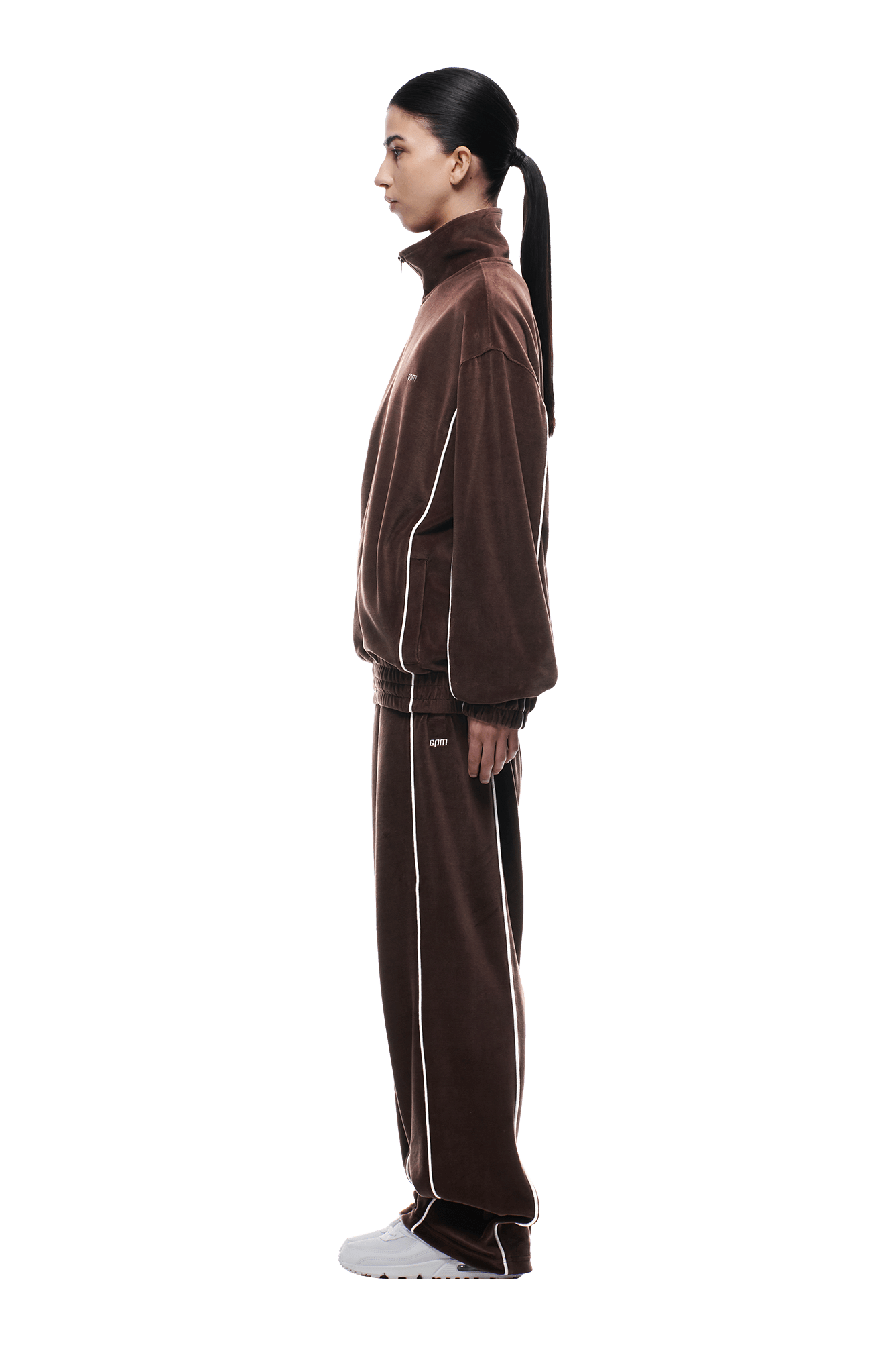 A person with long dark hair in a low ponytail stands in profile, wearing the 6PM VELOURS TRACK JACKET CHOCOLATE BROWN and white sneakers against a plain white background.