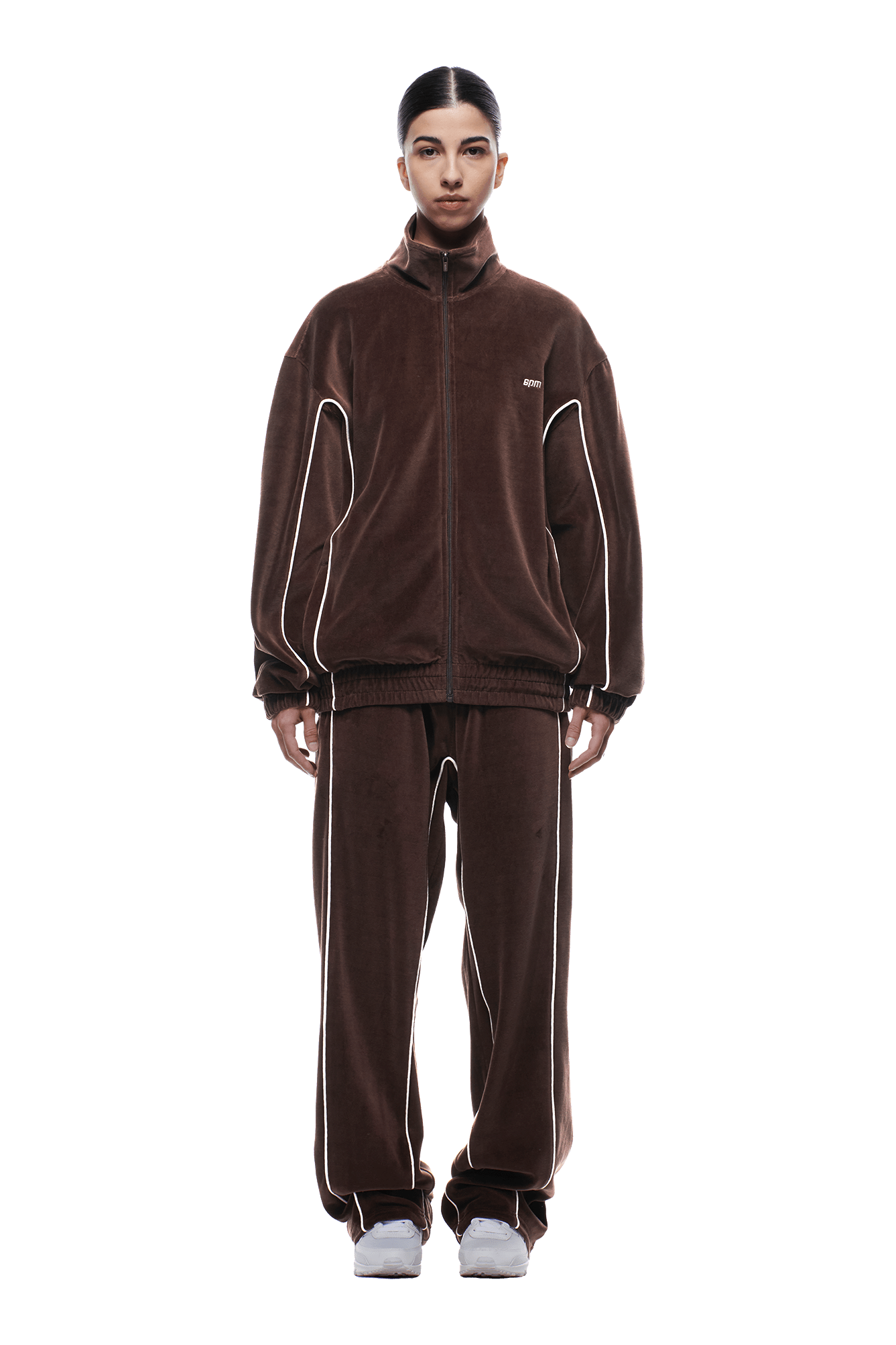 A person faces forward against a plain white background, wearing the 6PM VELOURS TRACK JACKET CHOCOLATE BROWN zipped up with matching relaxed-fit pants and white sneakers. The tracksuit features white piping for added detail.