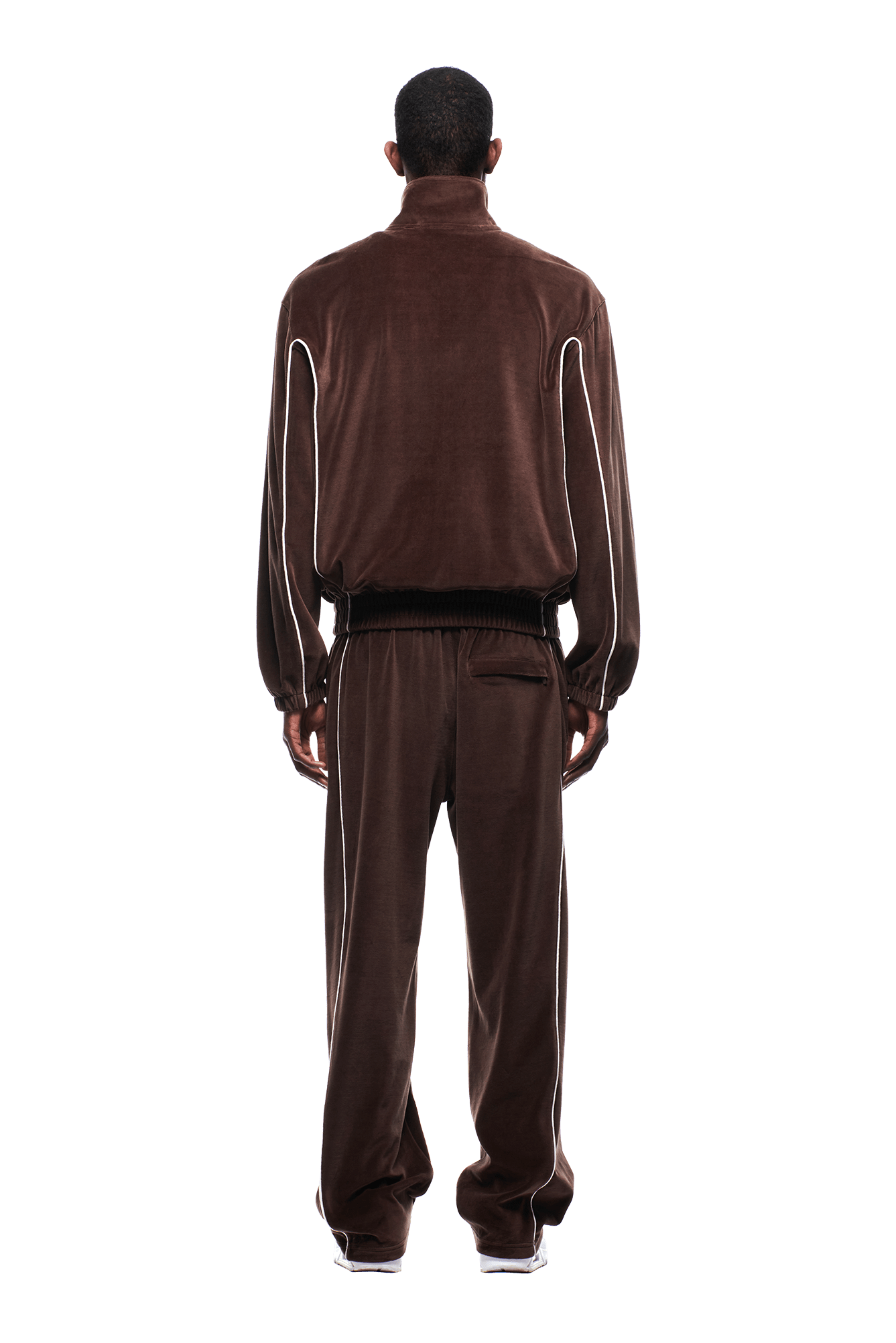 A person stands facing away on a white background, wearing the 6PM VELOURS TRACK JACKET CHOCOLATE BROWN with white piping details and white sneakers.