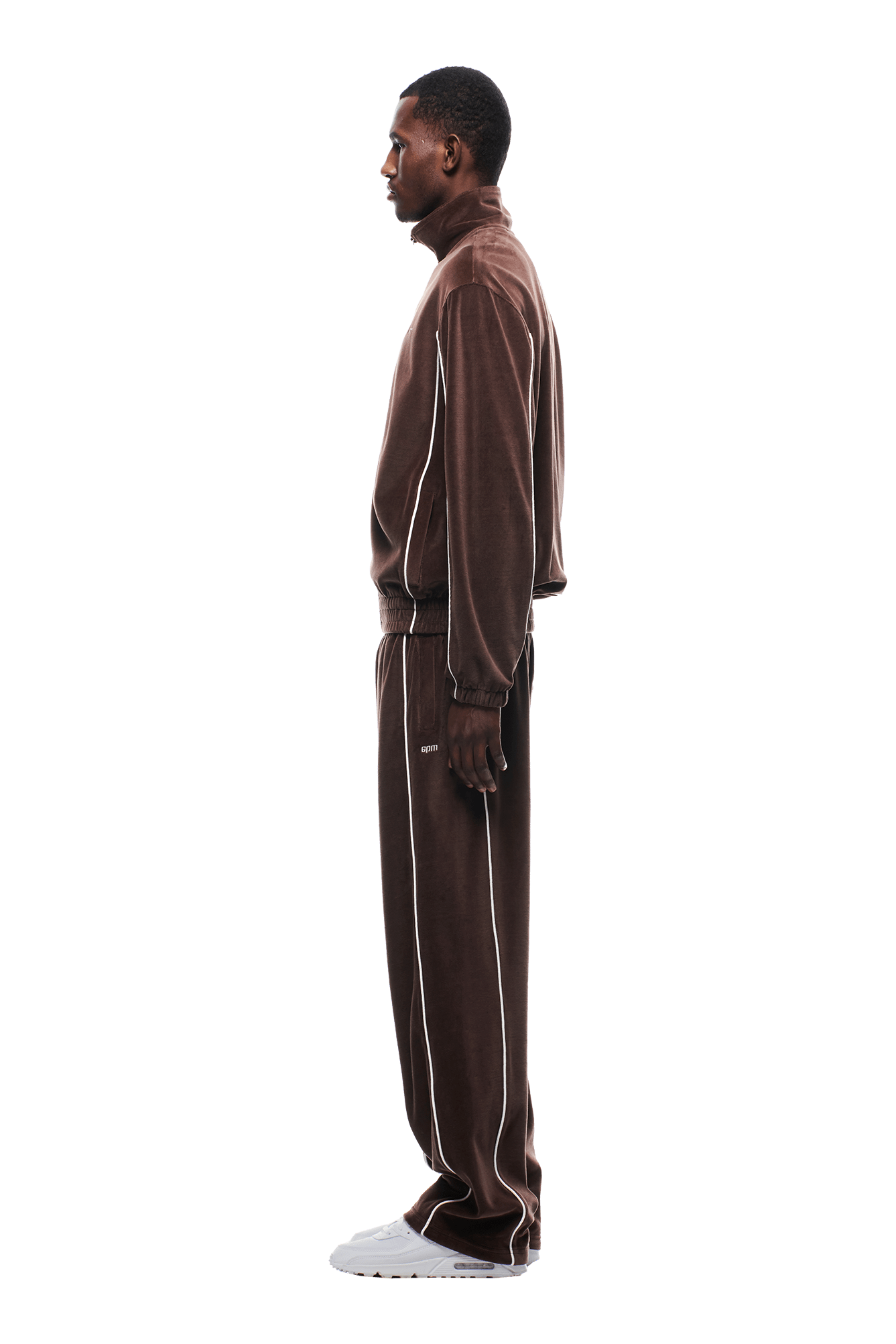 A man stands in profile against a plain white background, wearing the 6PM VELOURS TRACK JACKET CHOCOLATE BROWN with white side piping and white sneakers.