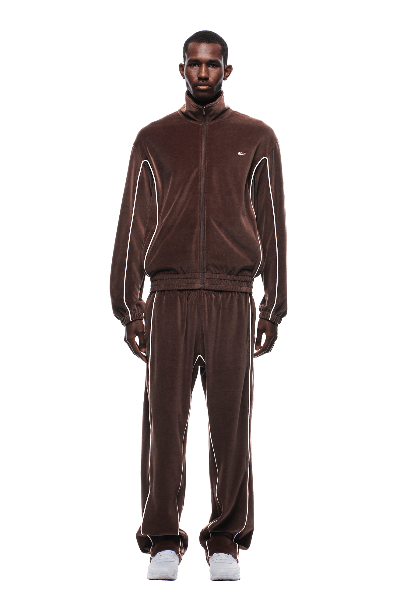 A man with short hair poses facing forward in a 6PM VELOURS TRACK JACKET CHOCOLATE BROWN with white piping, matching pants, and white sneakers, set against a plain white background.