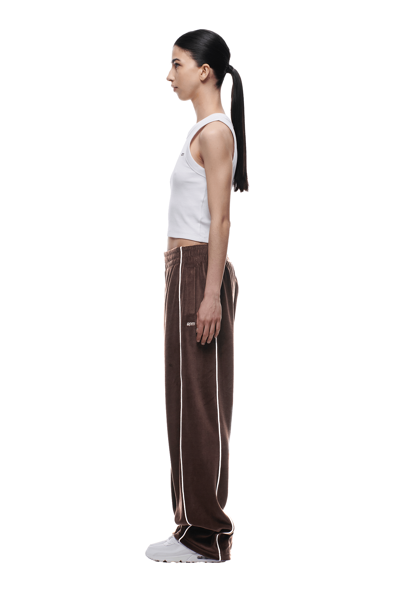A woman with long dark hair in a low ponytail stands in profile wearing the 6PM VELOURS TRACK PANT CHOCOLATE BROWN, a white sleeveless top, and white sneakers against a plain white background.