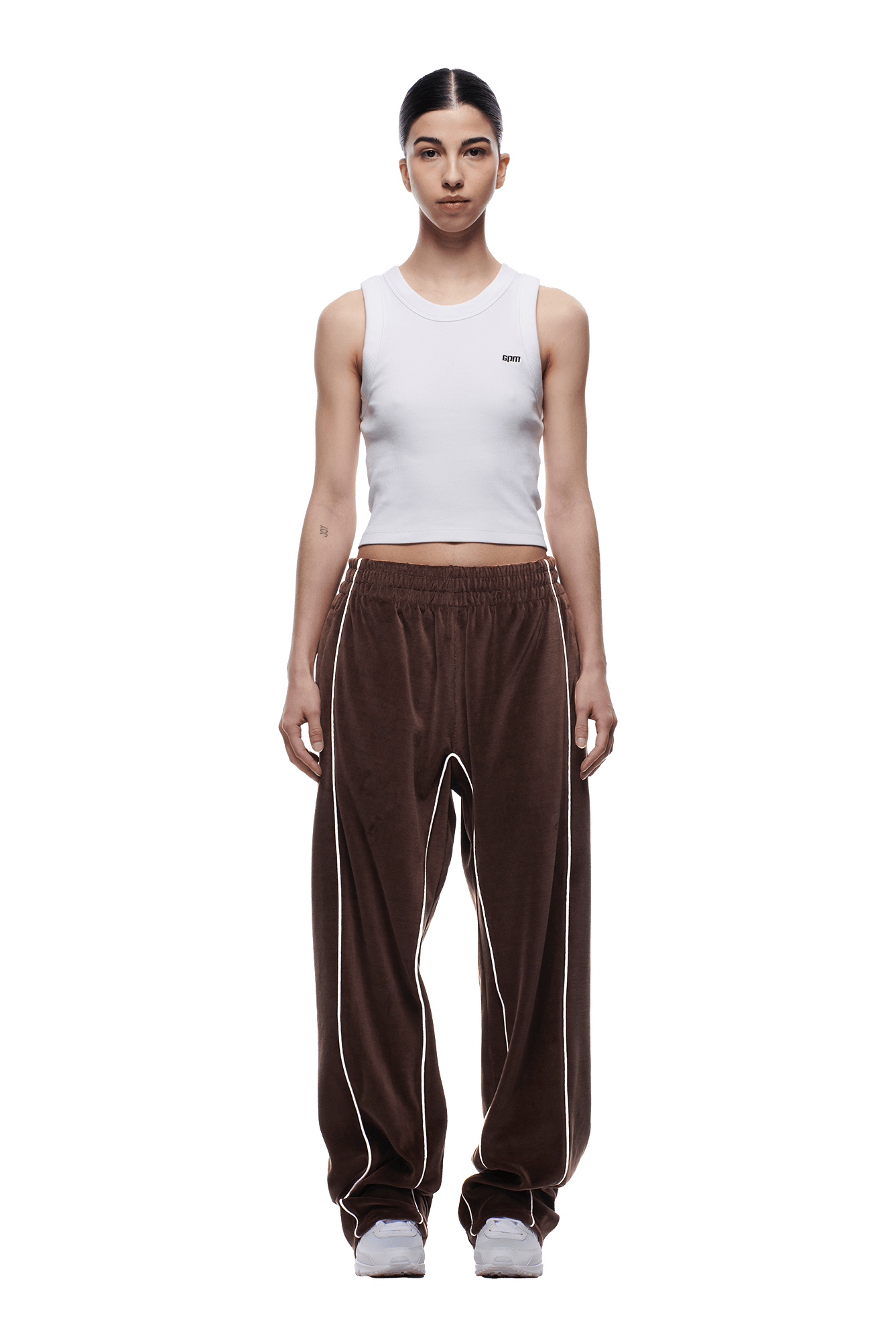 A person faces forward wearing the 6PM VELOURS TRACK PANT CHOCOLATE BROWN with white piping, paired with a white sleeveless crop top and gray sneakers against a plain white background.