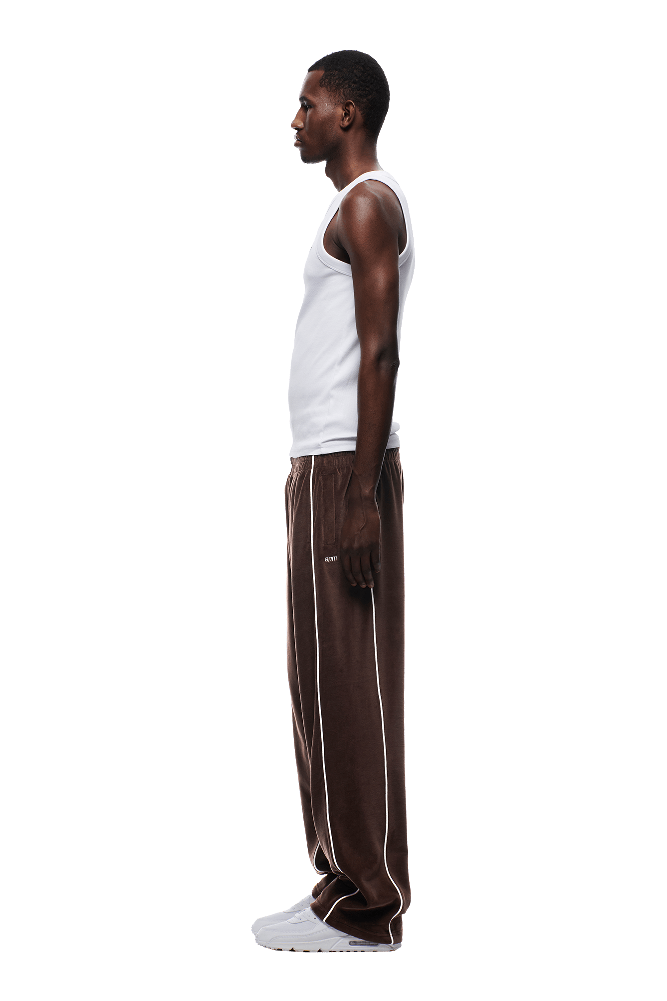 A man stands in profile against a white background, wearing 6PM’s VELOURS TRACK PANT CHOCOLATE BROWN with white piping, paired with a white tank top and white sneakers. His arms are relaxed at his sides.