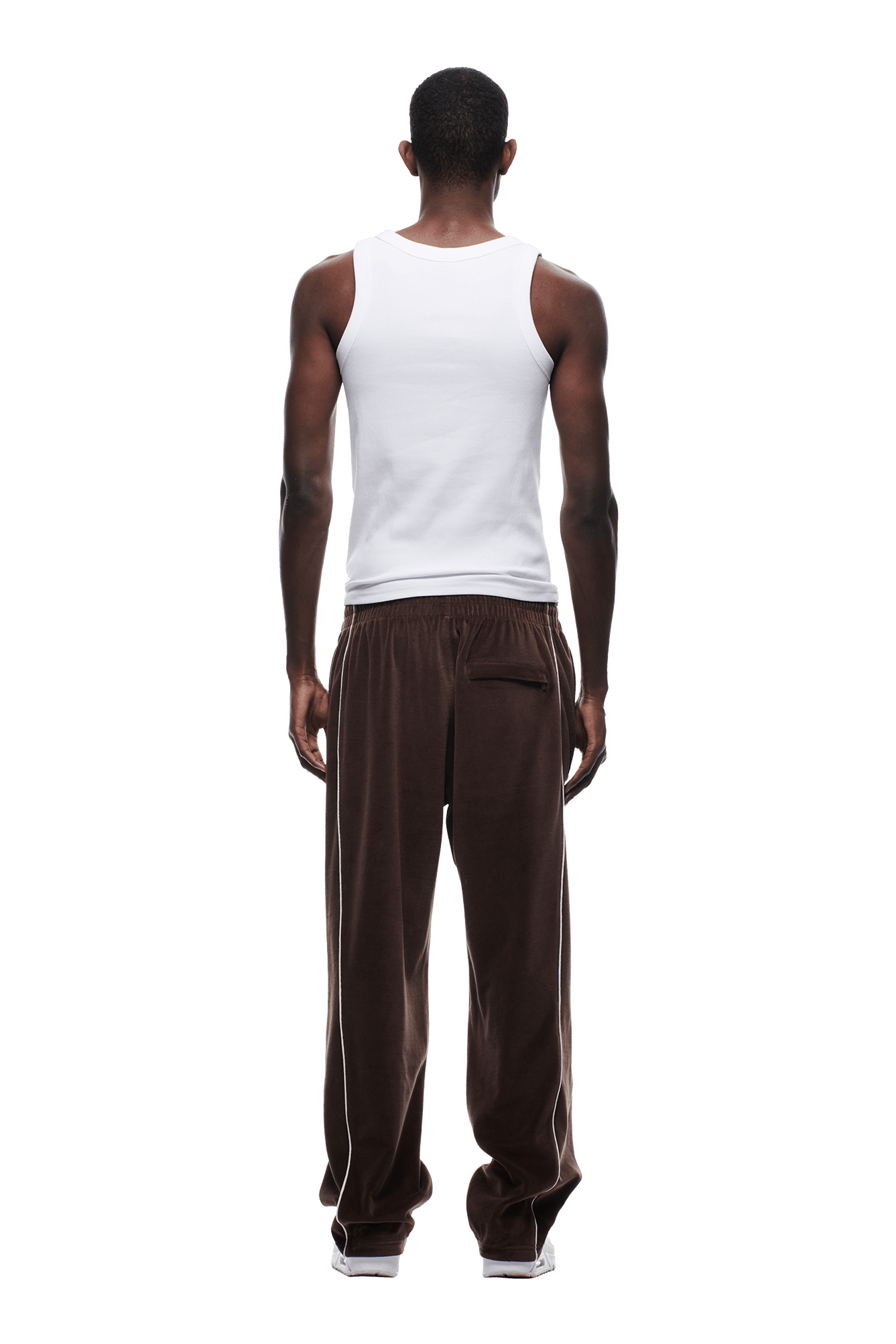 A man, seen from behind, wears the 6PM VELOURS TRACK PANT CHOCOLATE BROWN—loose brown sweatpants with white piping—paired with a white sleeveless tank top, standing against a plain white background.