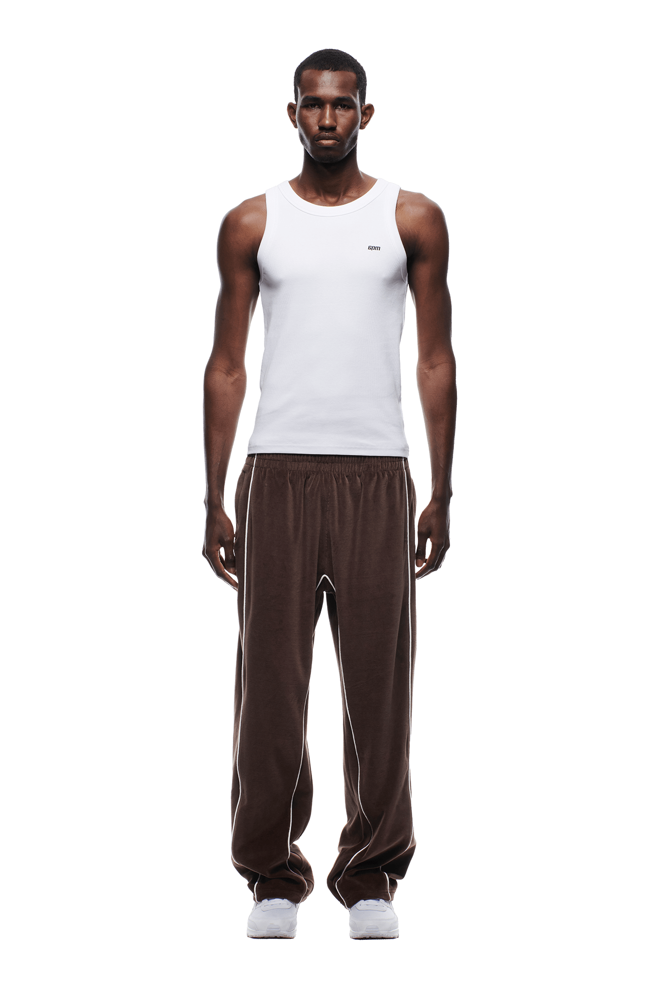 A man stands facing forward against a plain white background, wearing the 6PM VELOURS TRACK PANT CHOCOLATE BROWN, a white sleeveless tank top, and white sneakers.