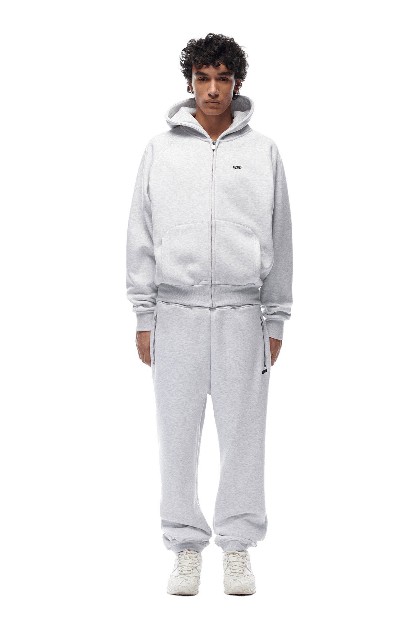 A person faces forward in the 6PM ZIP HOODIE LIGHT GREY with matching sweatpants and white sneakers, highlighting a clean look ideal for daily wear, set against a transparent background.
