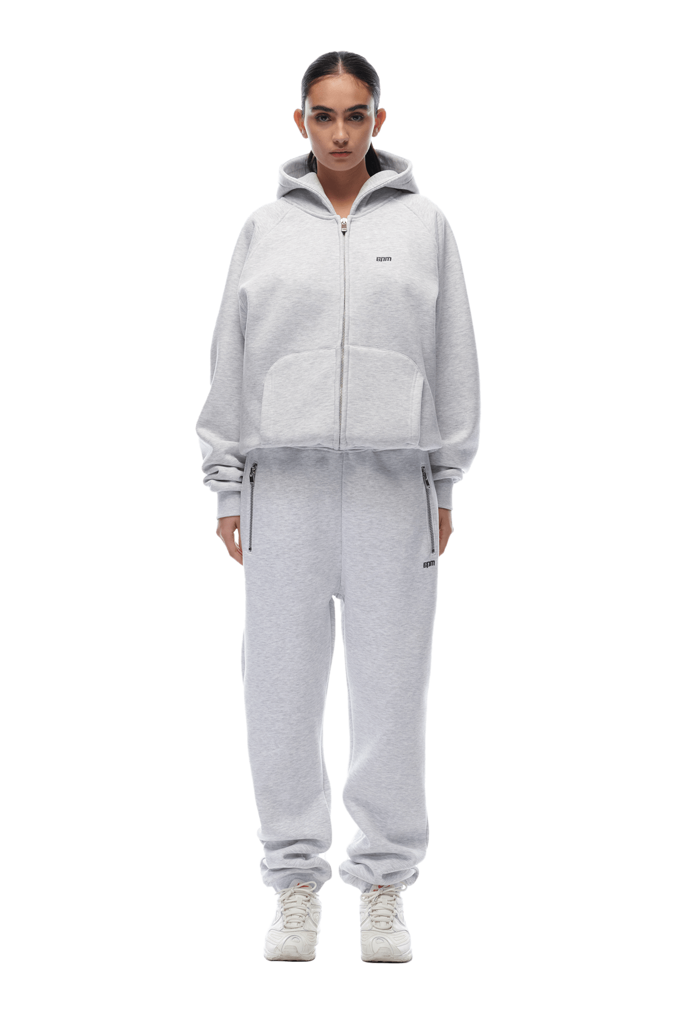 A person stands facing forward, wearing the 6PM ZIP HOODIE LIGHT GREY and matching sweatpants with white sneakers. The casual, sporty look is ideal for everyday wear. Their hands rest by their sides against a transparent background.