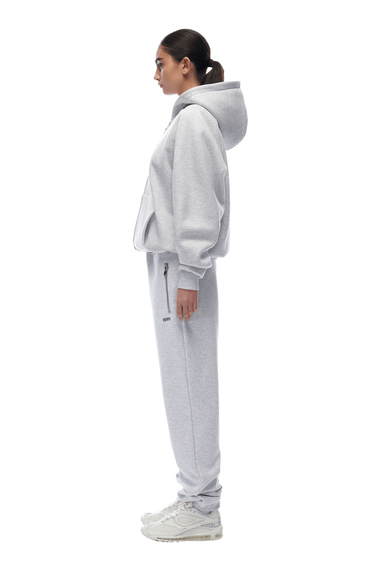 A woman stands in profile wearing the 6PM ZIP HOODIE LIGHT GREY and matching sweatpants with white sneakers. Her hair is tied back as she faces left against a transparent background, showcasing the outfit’s clean, everyday style.