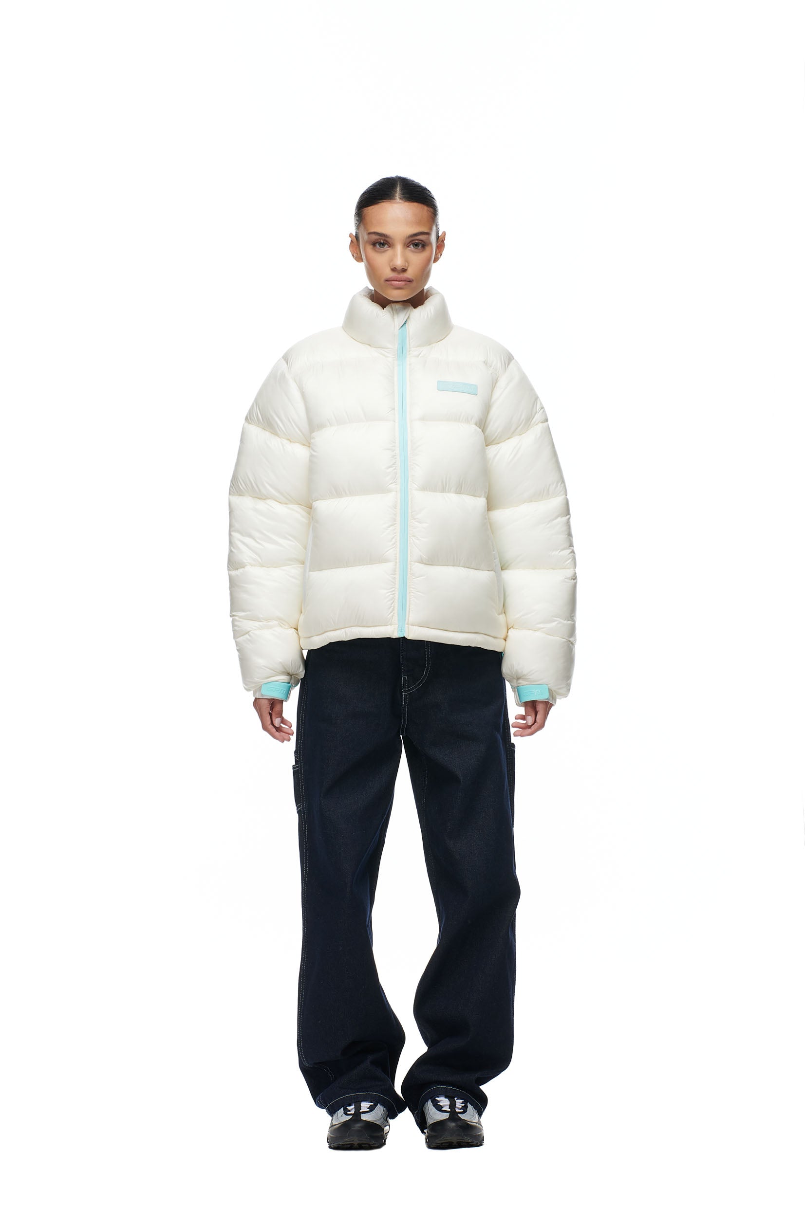 A person models the 6PM PUFFIEST PUFFER COCONUT WHITE jacket, paired with dark blue jeans and black sneakers, standing forward against a plain white background.