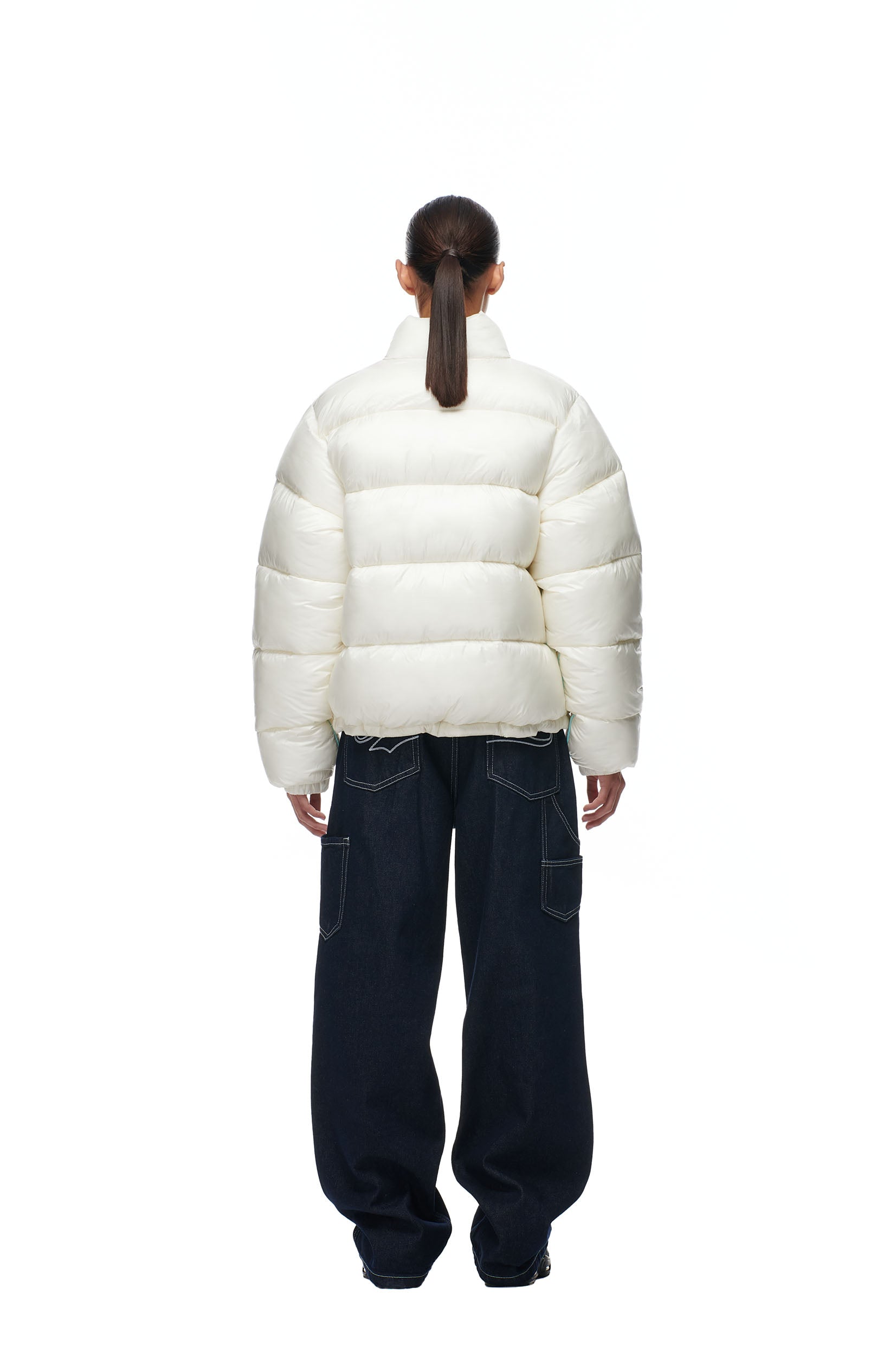 A person with a ponytail, seen from behind, wears the 6PM PUFFIEST PUFFER COCONUT WHITE jacket and loose dark jeans with visible stitching, standing against a plain white background.