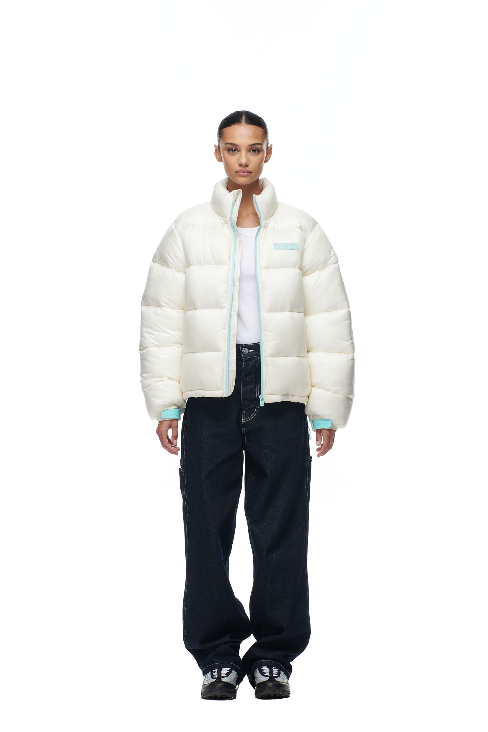 A person stands facing forward in the 6PM PUFFIEST PUFFER COCONUT WHITE jacket, a white top, loose dark jeans, and black sneakers against a plain white background.