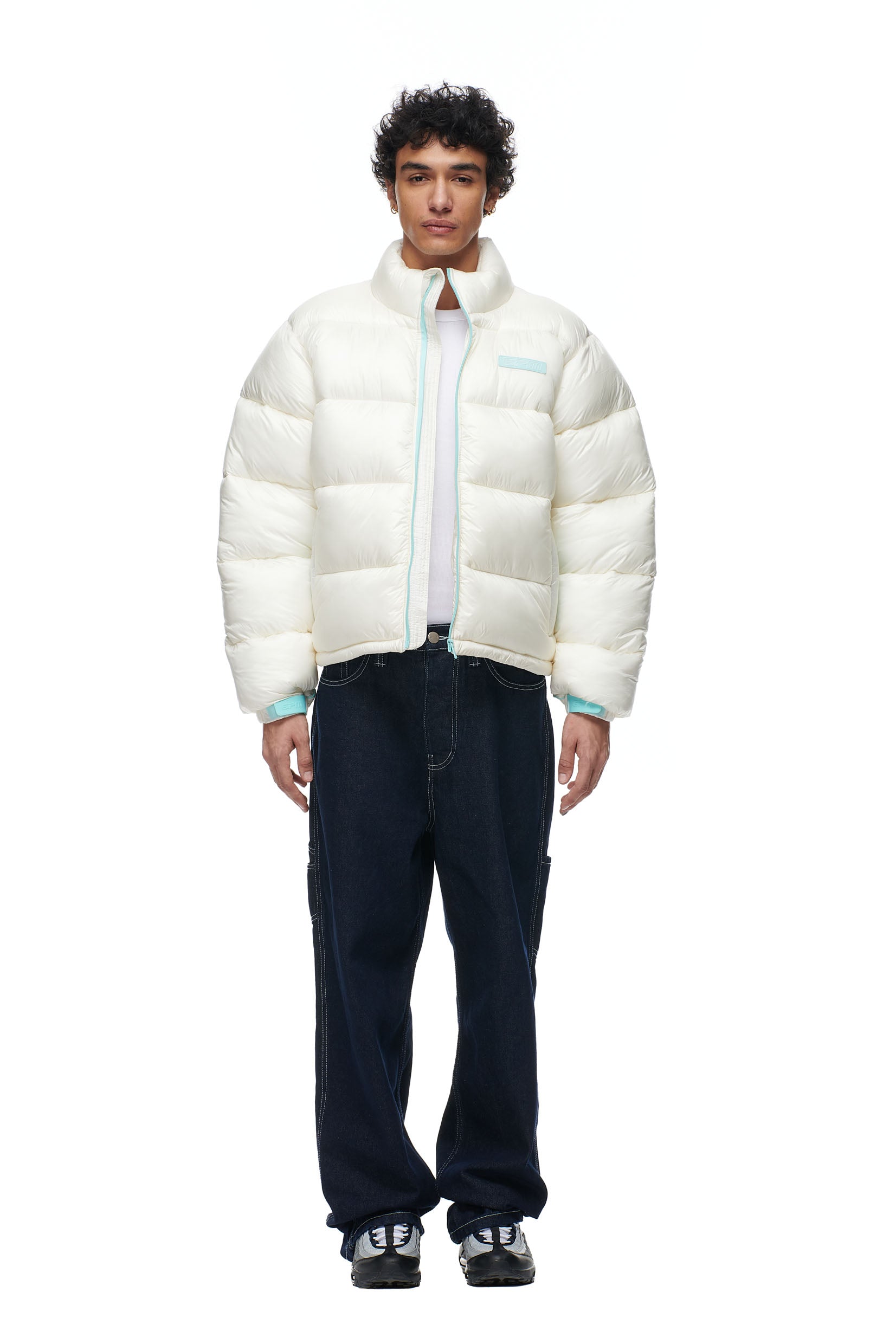 A person faces forward against a plain white background wearing the 6PM PUFFIEST PUFFER COCONUT WHITE jacket, paired with dark blue baggy jeans and black sneakers.