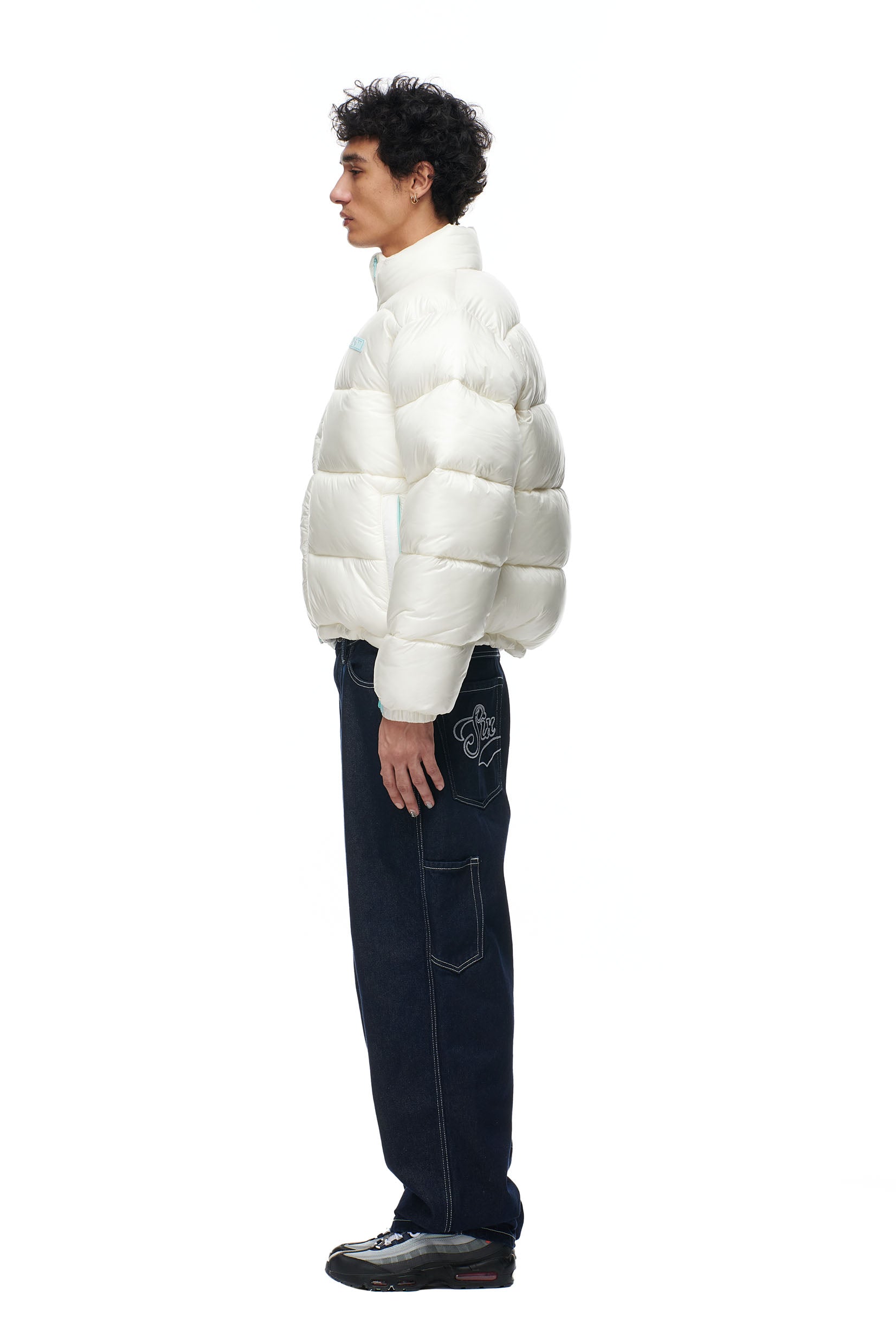 A person stands in profile against a plain white background, wearing the 6PM PUFFIEST PUFFER COCONUT WHITE jacket, dark blue jeans with pocket embroidery, and black sneakers.