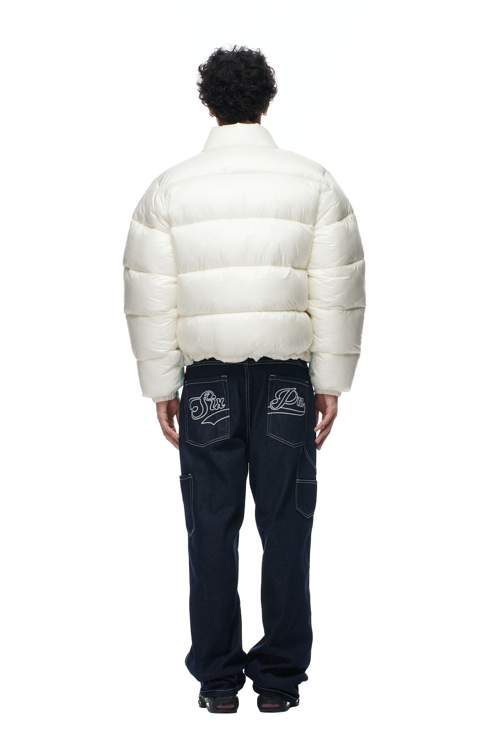 A person faces away from the camera wearing the 6PM PUFFIEST PUFFER COCONUT WHITE jacket and dark blue jeans with white embroidery on the back pockets, set against a plain white background.