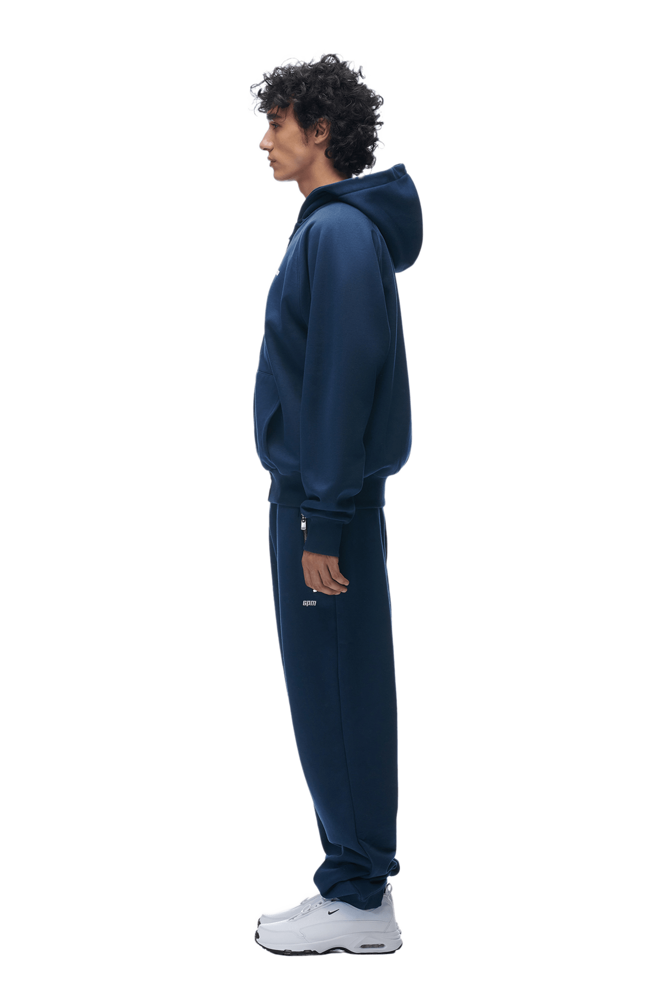 A person with curly hair stands in profile, wearing the 6PM ZIP HOODIE NAVY and matching sweatpants for a structured, clean look with white sneakers, set against a plain background.