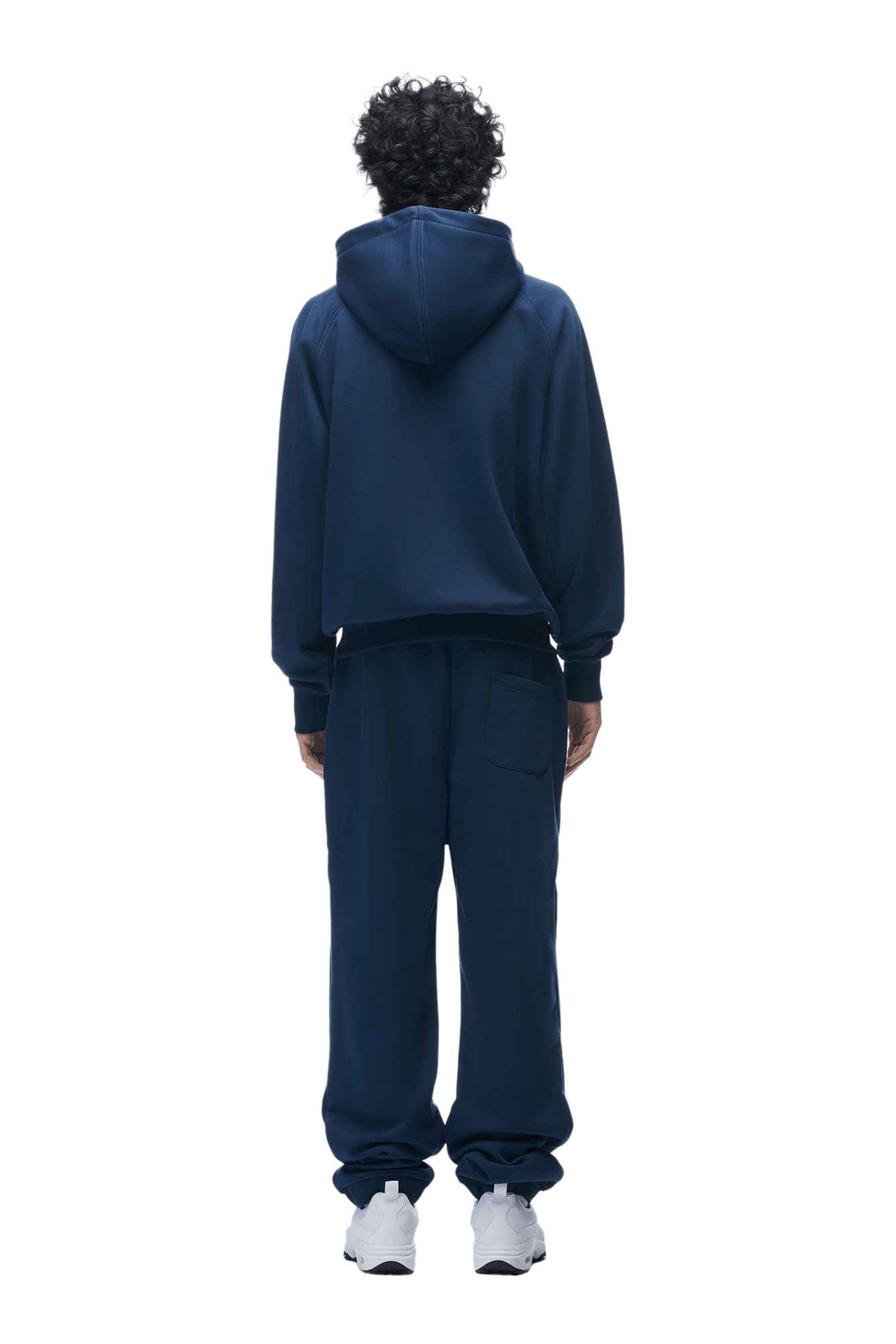 A person with curly hair, facing away, wears the 6PM ZIP HOODIE NAVY and matching sweatpants with white sneakers. The outfit is clean and versatile for everyday wear, shown against a plain transparent background.