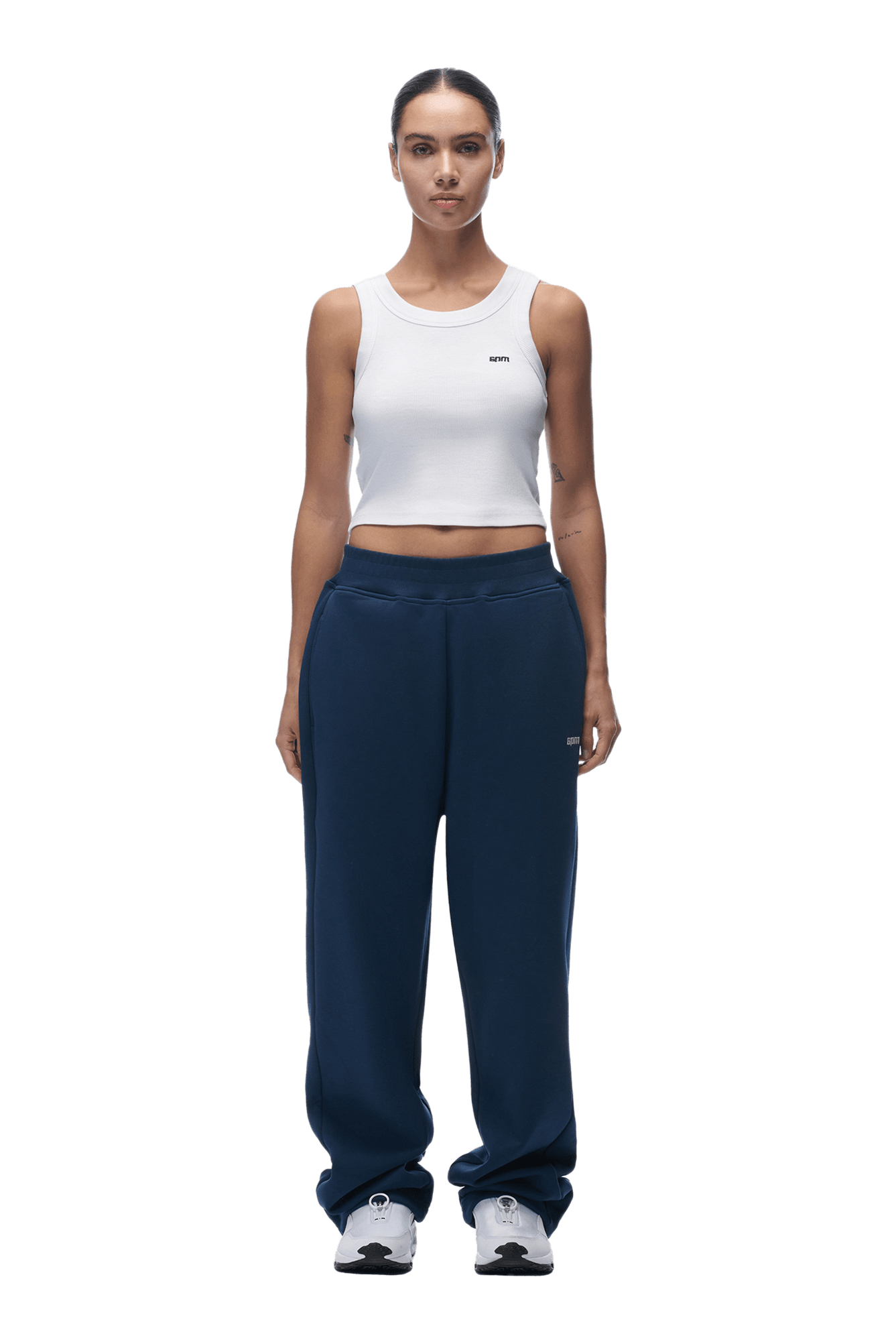A person stands facing forward in a white sleeveless crop top and 6PM OPEN LEG JOGGER NAVY sweatpants with perfect stacking length, styled with white sneakers. Their hair is pulled back tightly, expression neutral, on a transparent background.