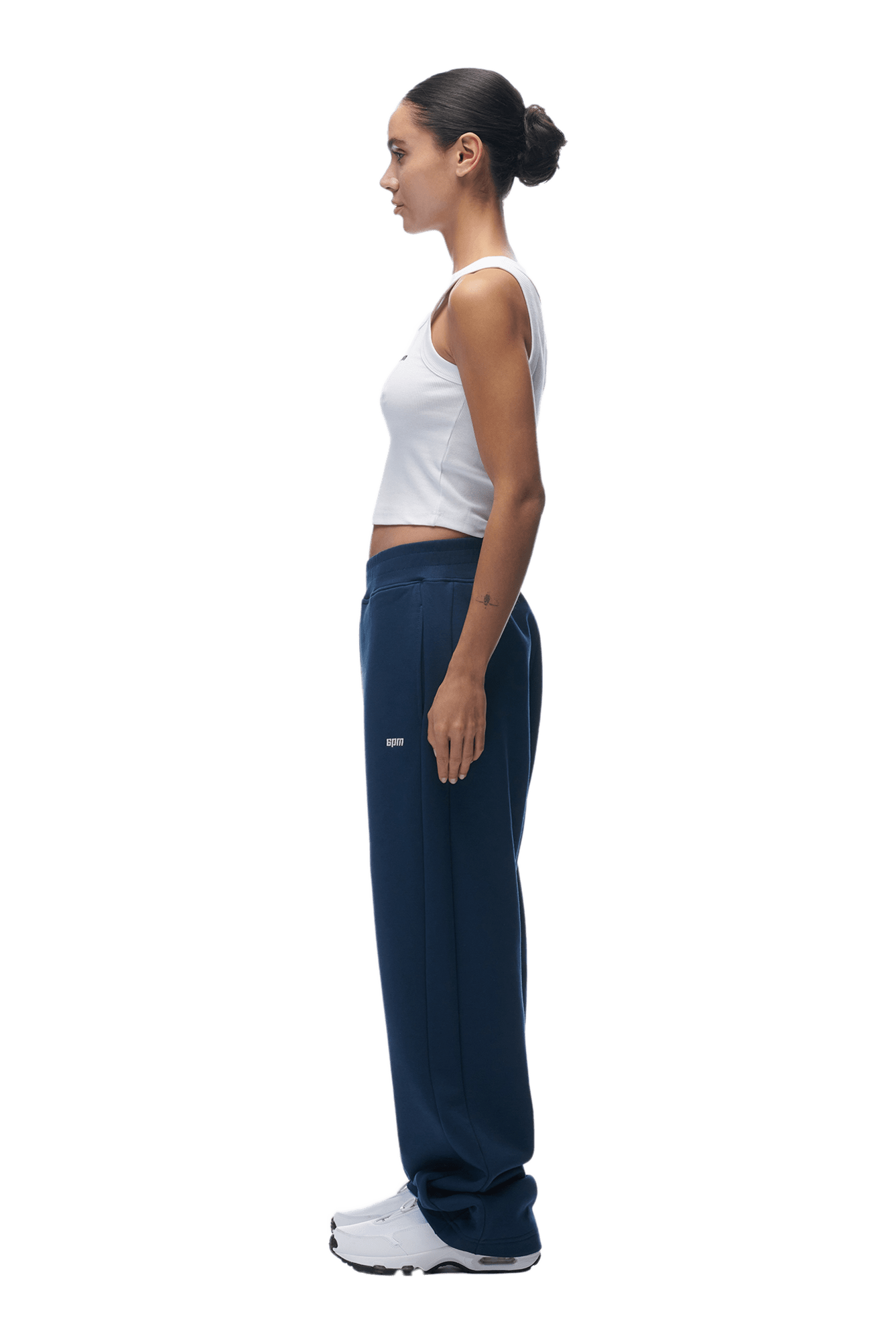 A woman stands in profile facing left, wearing a white tank top, 6PM OPEN LEG JOGGER NAVY sweatpants with perfect stacking length, and white sneakers. Her hair is tied in a bun, arms at her sides. The background is transparent.