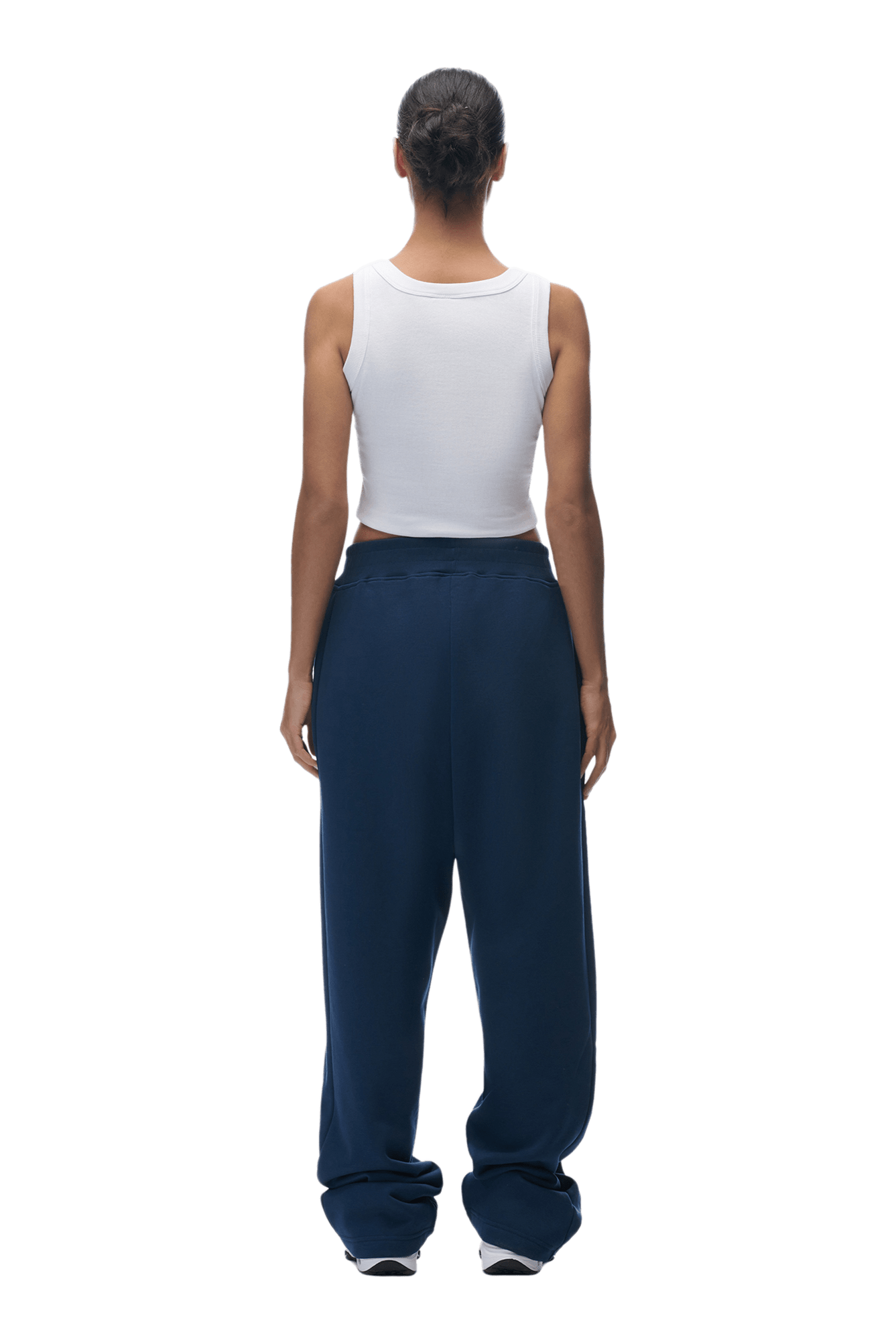 A person with dark hair in a low bun faces away, wearing a white sleeveless tank top and 6PM OPEN LEG JOGGER NAVY pants with the perfect stacking length, paired with white sneakers, against a plain background.