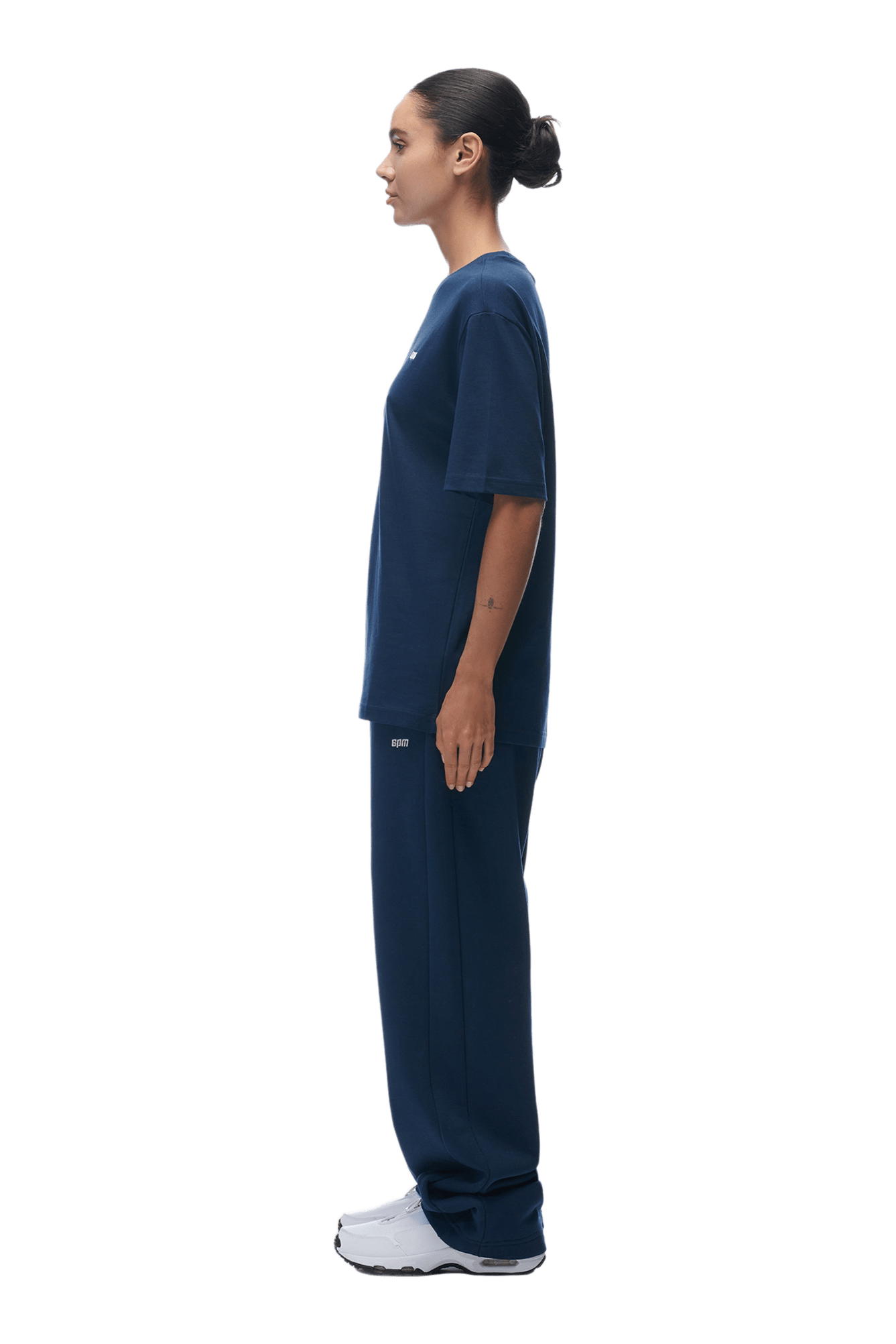 A woman in profile, facing left, wears the 6PM T-SHIRT NAVY with matching pants and white sneakers. Her hair is tied in a neat bun. The background is transparent.