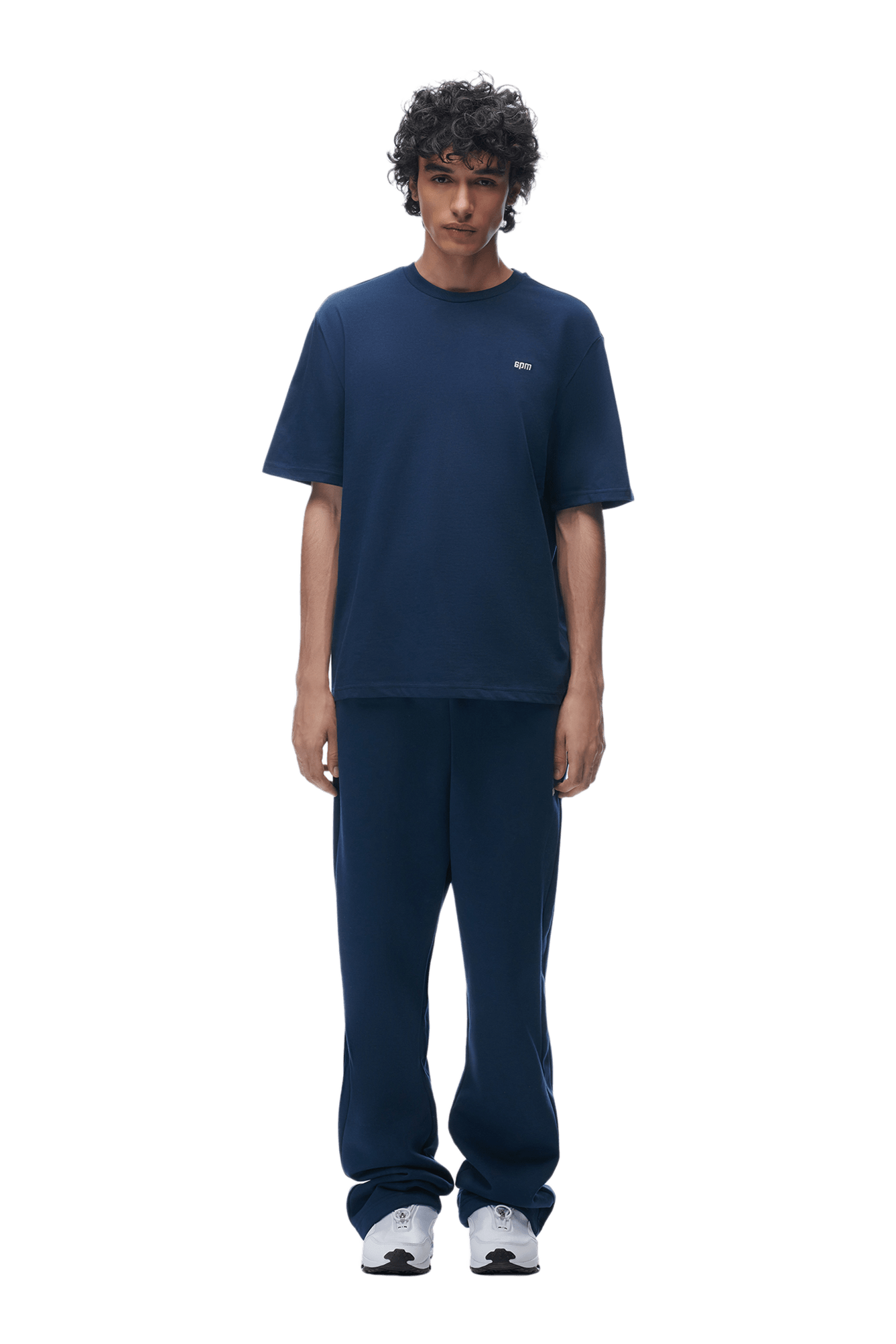 A person with curly hair stands facing forward, wearing the 6PM T-SHIRT NAVY and matching pants with white sneakers, against a plain background.