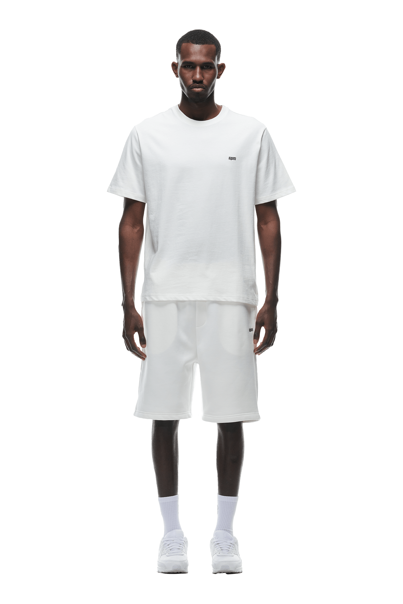 A man stands facing forward in a 6PM T-SHIRT COCONUT WHITE, knee-length white shorts, white socks, and white sneakers against a solid green background.