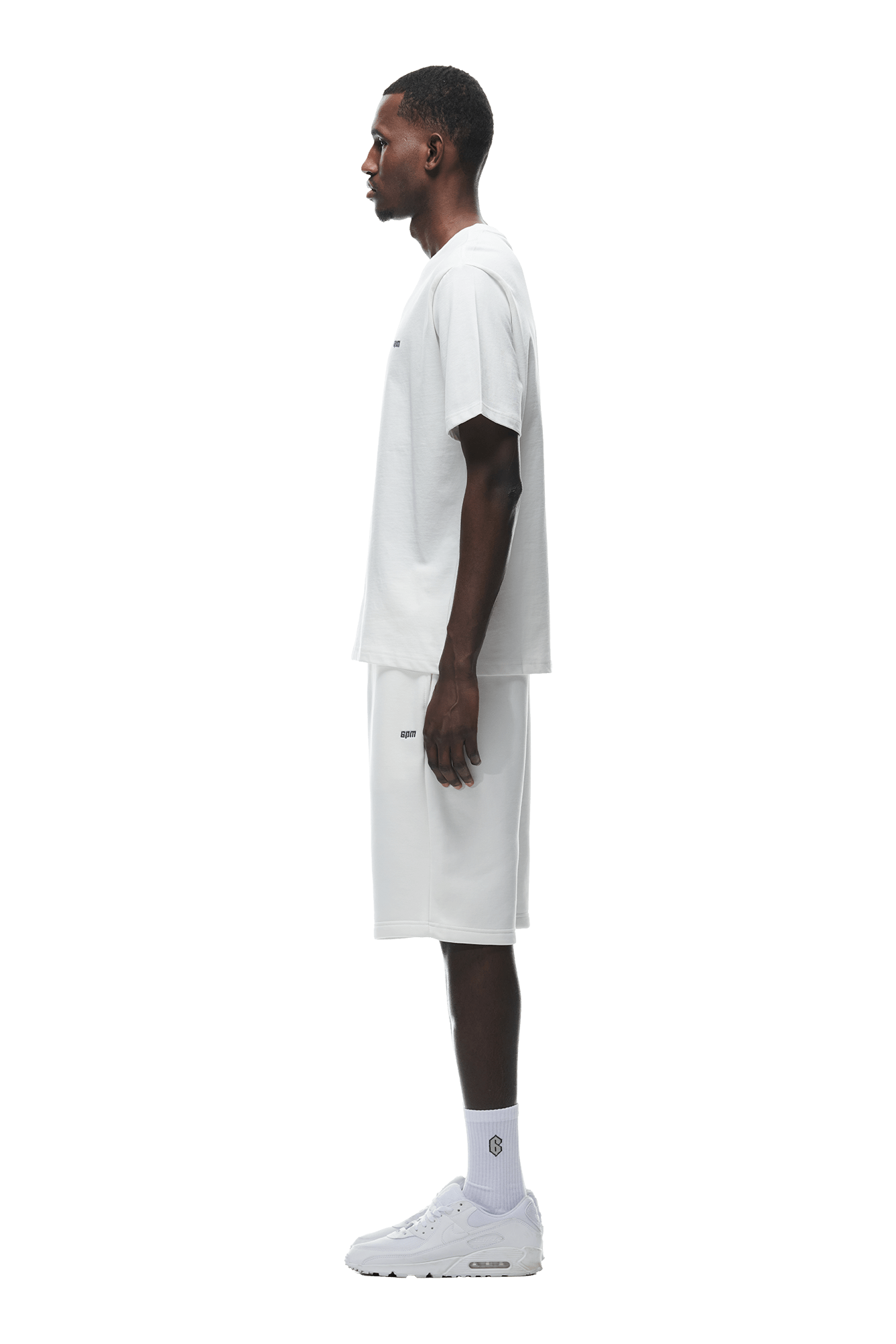 A man stands in profile facing left, wearing the 6PM T-SHIRT COCONUT WHITE, white knee-length shorts, white socks, and white sneakers against a plain background.