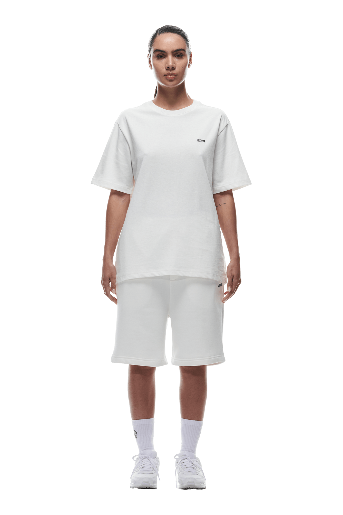 A person faces forward against a green background, wearing the 6PM T-SHIRT COCONUT WHITE, white knee-length shorts, white socks, and white sneakers.