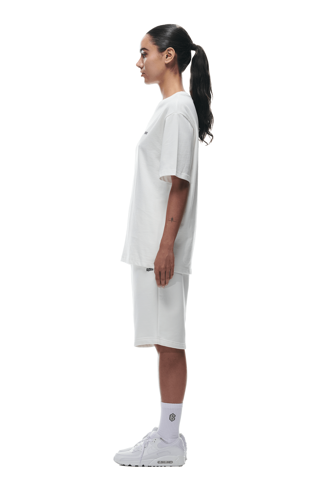A woman with dark hair in a ponytail stands in profile, wearing the 6PM T-SHIRT COCONUT WHITE, white knee-length shorts, socks, and sneakers against a transparent background.