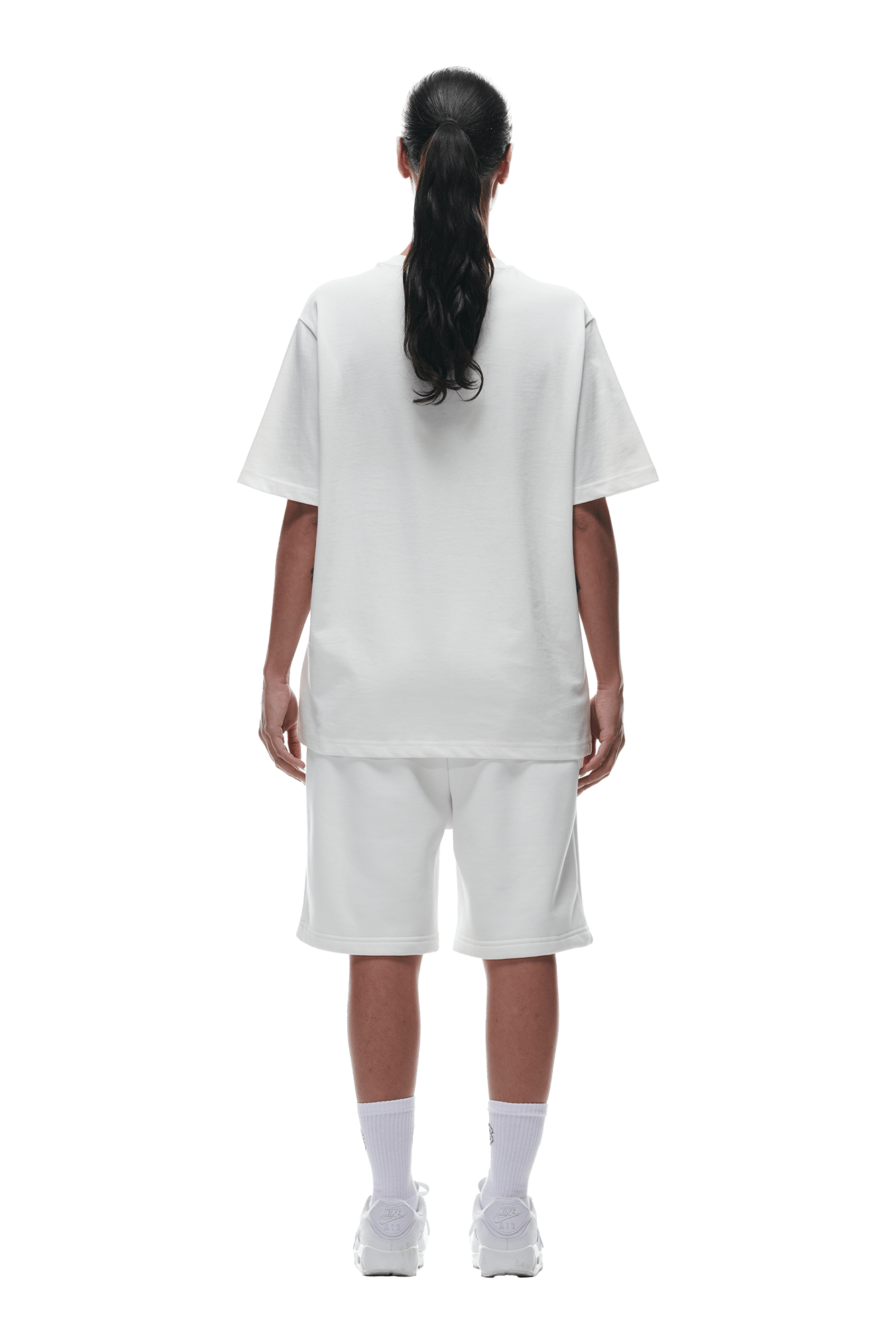 A person with long dark hair in a ponytail faces away, wearing the 6PM T-SHIRT COCONUT WHITE, white knee-length shorts, white socks, and sneakers, against a transparent background.
