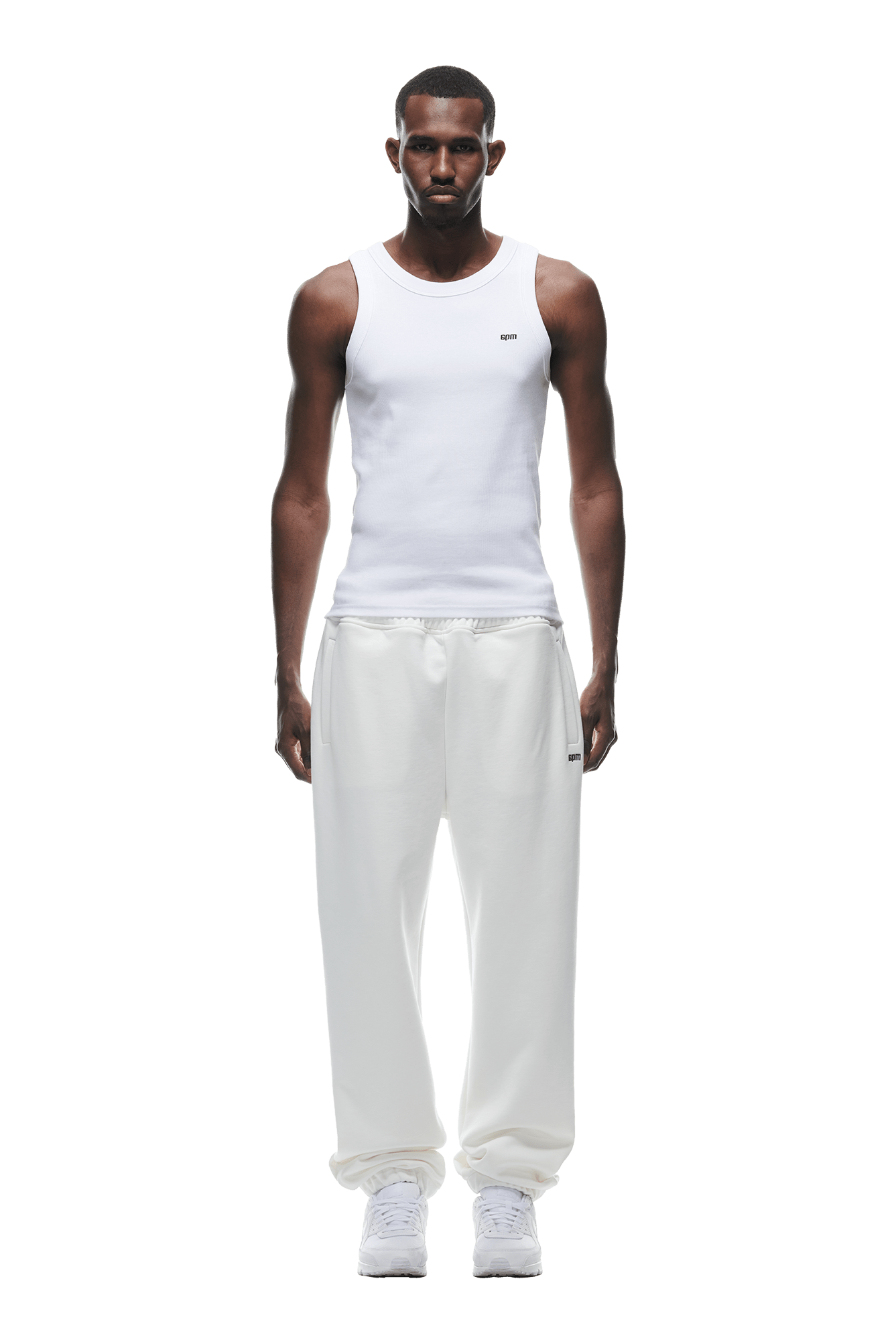 A man faces forward, wearing a white sleeveless tank, 6PM CUFF JOGGER COCONUT WHITE sweatpants with cuffed legs, and white sneakers. The background is transparent.