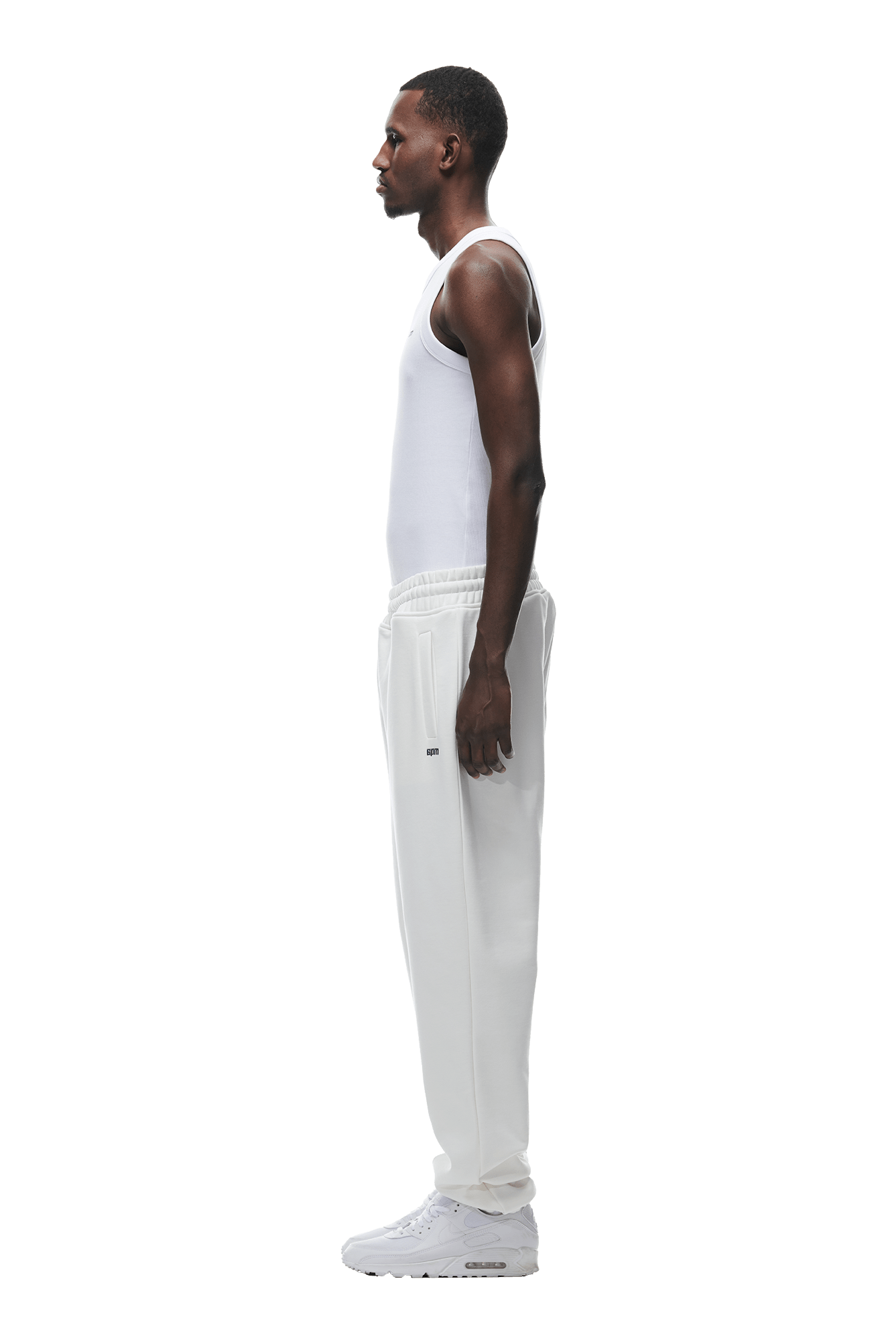 A man in profile faces left, wearing a 6PM CUFF JOGGER COCONUT WHITE sweatpants and a white tank top with white sneakers, set against a plain green background.