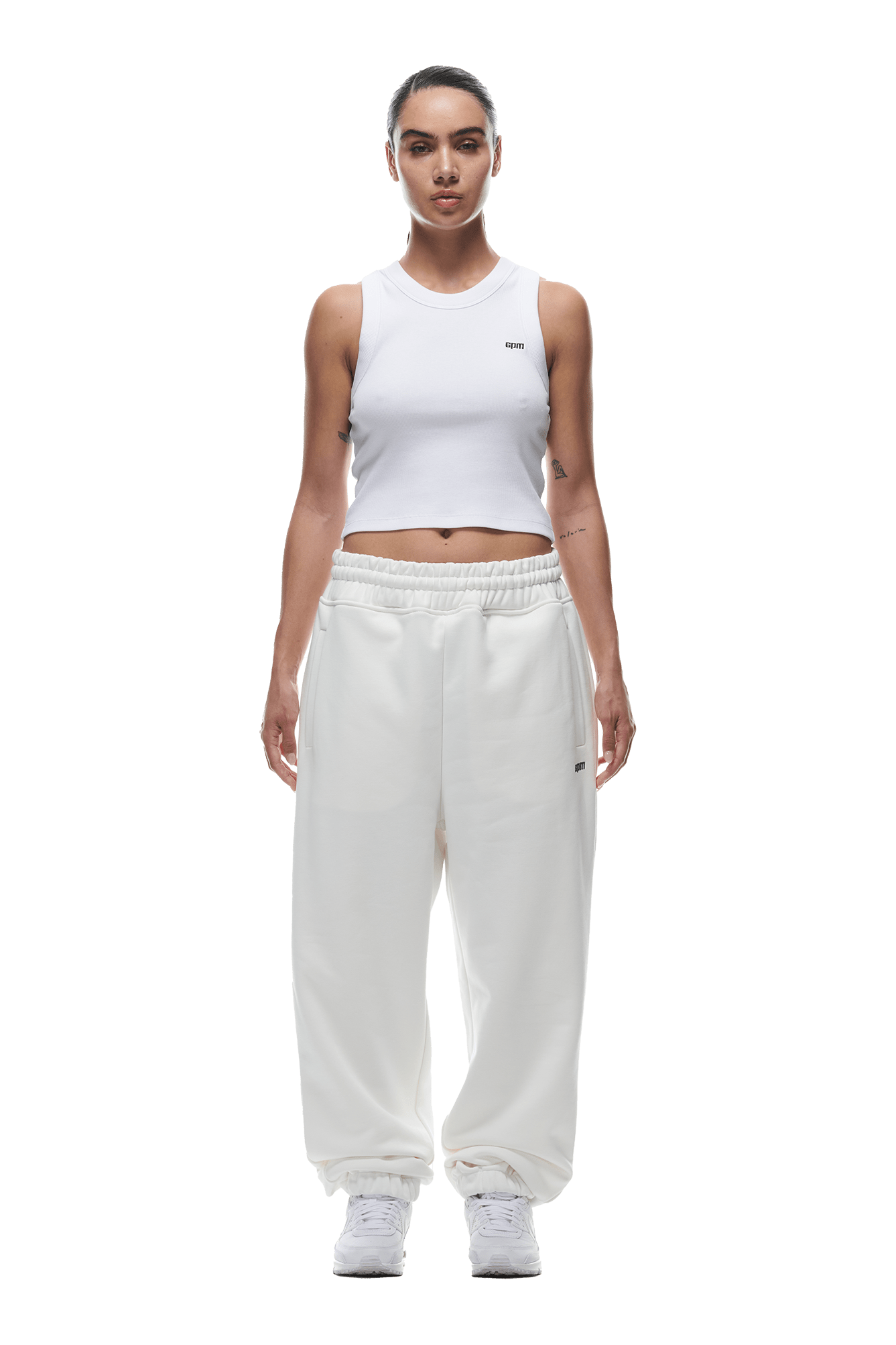 A person stands facing forward in a white sleeveless crop top and 6PM CUFF JOGGER COCONUT WHITE sweatpants with cuffed legs, paired with white sneakers. Their hair is pulled back and arms relaxed at their sides, against a plain background.
