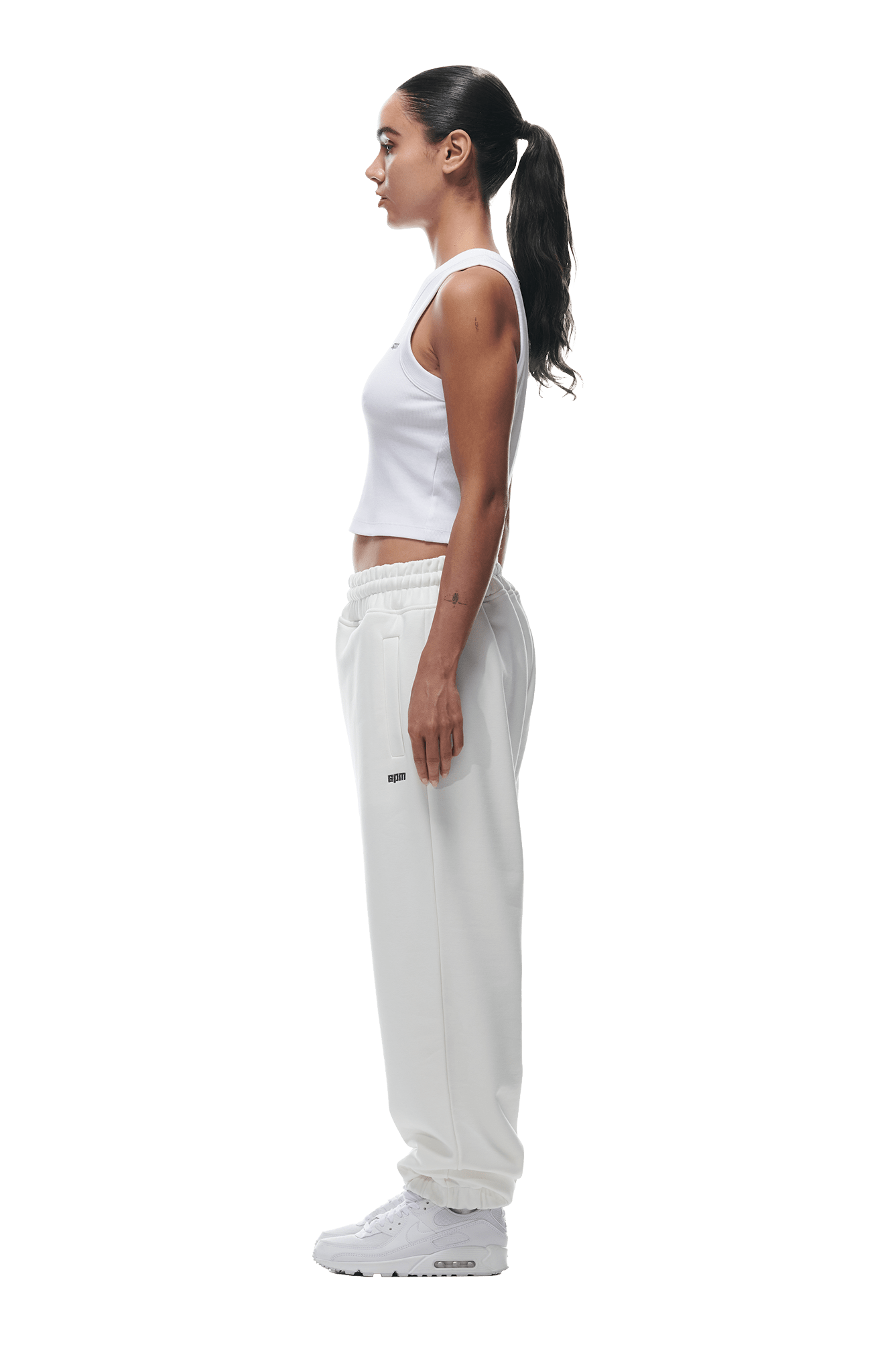 A woman with long dark hair in a ponytail stands in profile wearing a white sleeveless top, 6PM CUFF JOGGER COCONUT WHITE sweatpants, and white sneakers against a plain background.