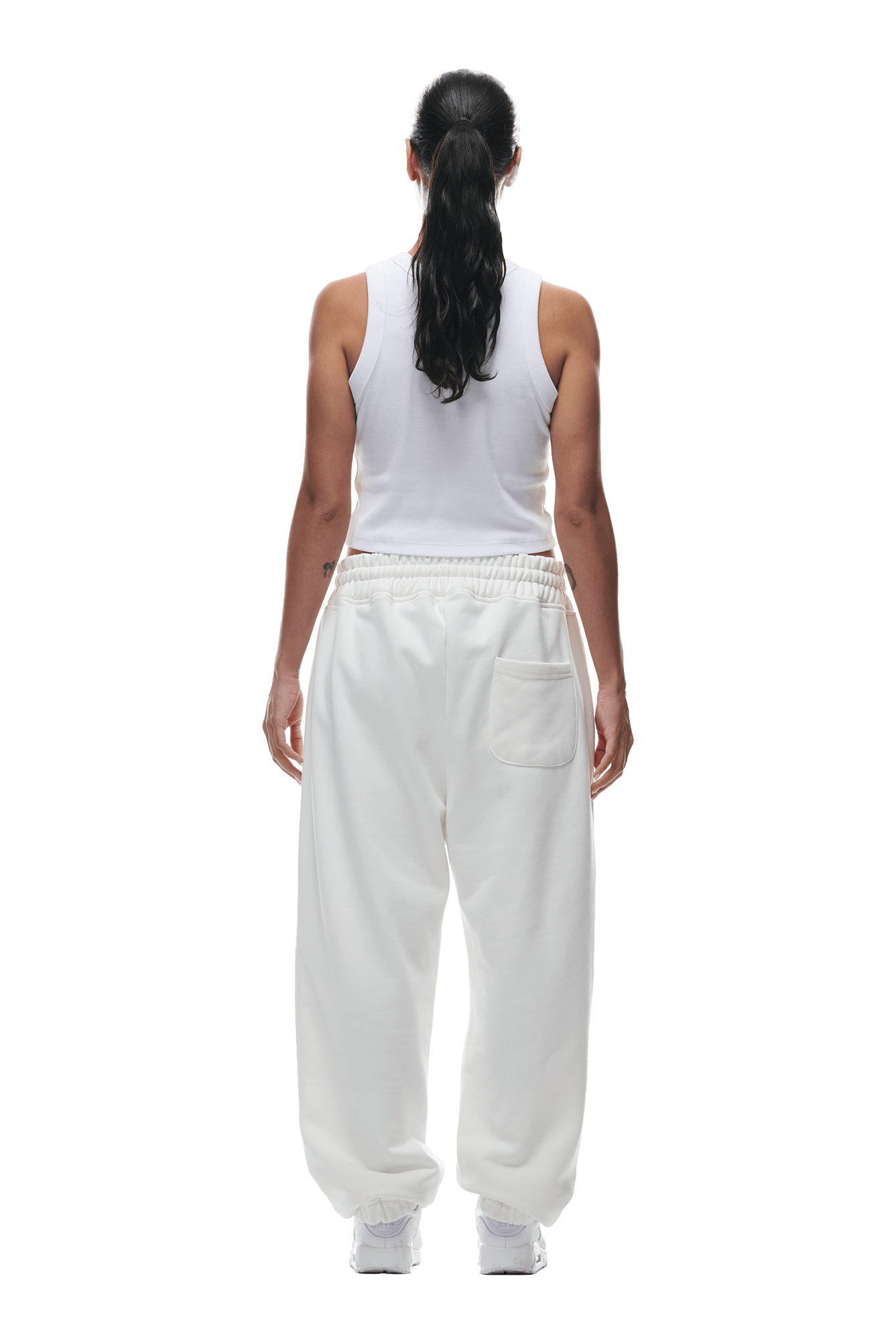 A person with long dark hair in a ponytail stands facing away, wearing 6PM CUFF JOGGER COCONUT WHITE sweatpants and a white sleeveless top with white sneakers, set against a plain background.