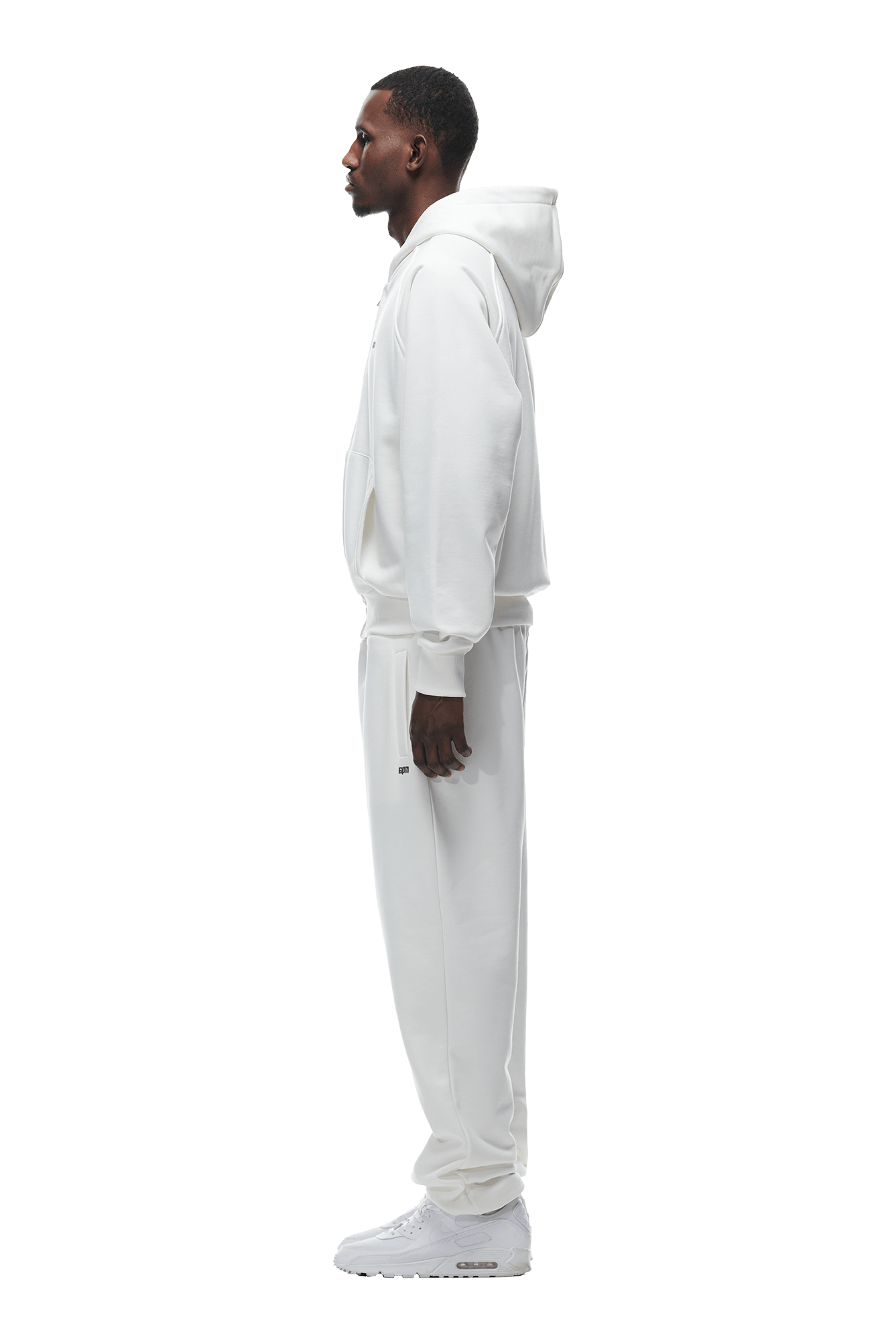 A man stands in profile facing left, wearing the 6PM ZIP HOODIE COCONUT WHITE with matching sweatpants and sneakers against a plain backdrop—a clean, versatile look ideal for everyday wear.