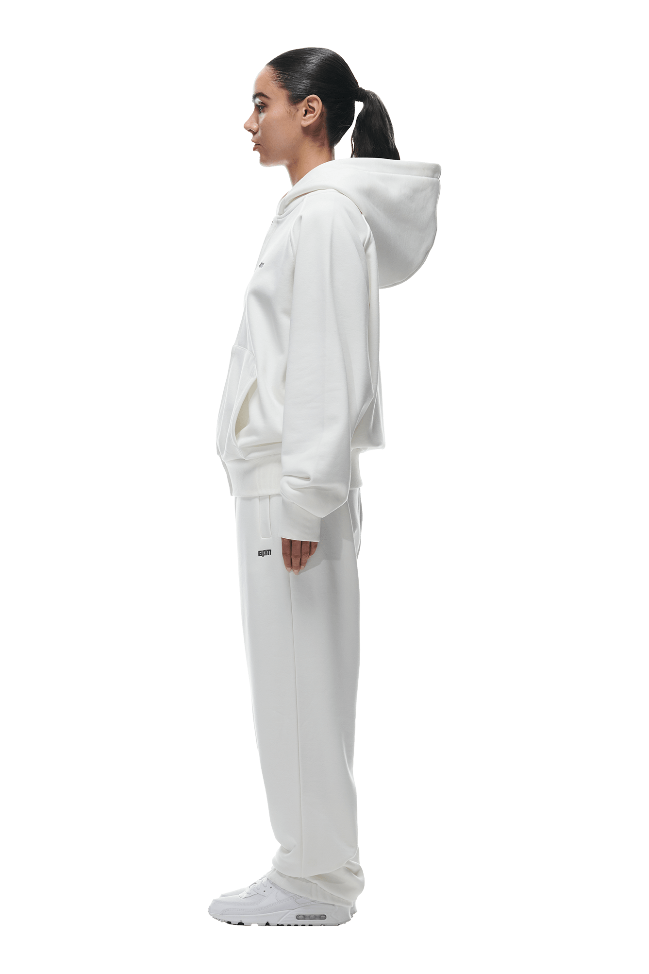 A woman with a ponytail stands in profile wearing the 6PM ZIP HOODIE COCONUT WHITE, paired with matching sweatpants and sneakers—an effortlessly clean look for everyday wear against a plain background.